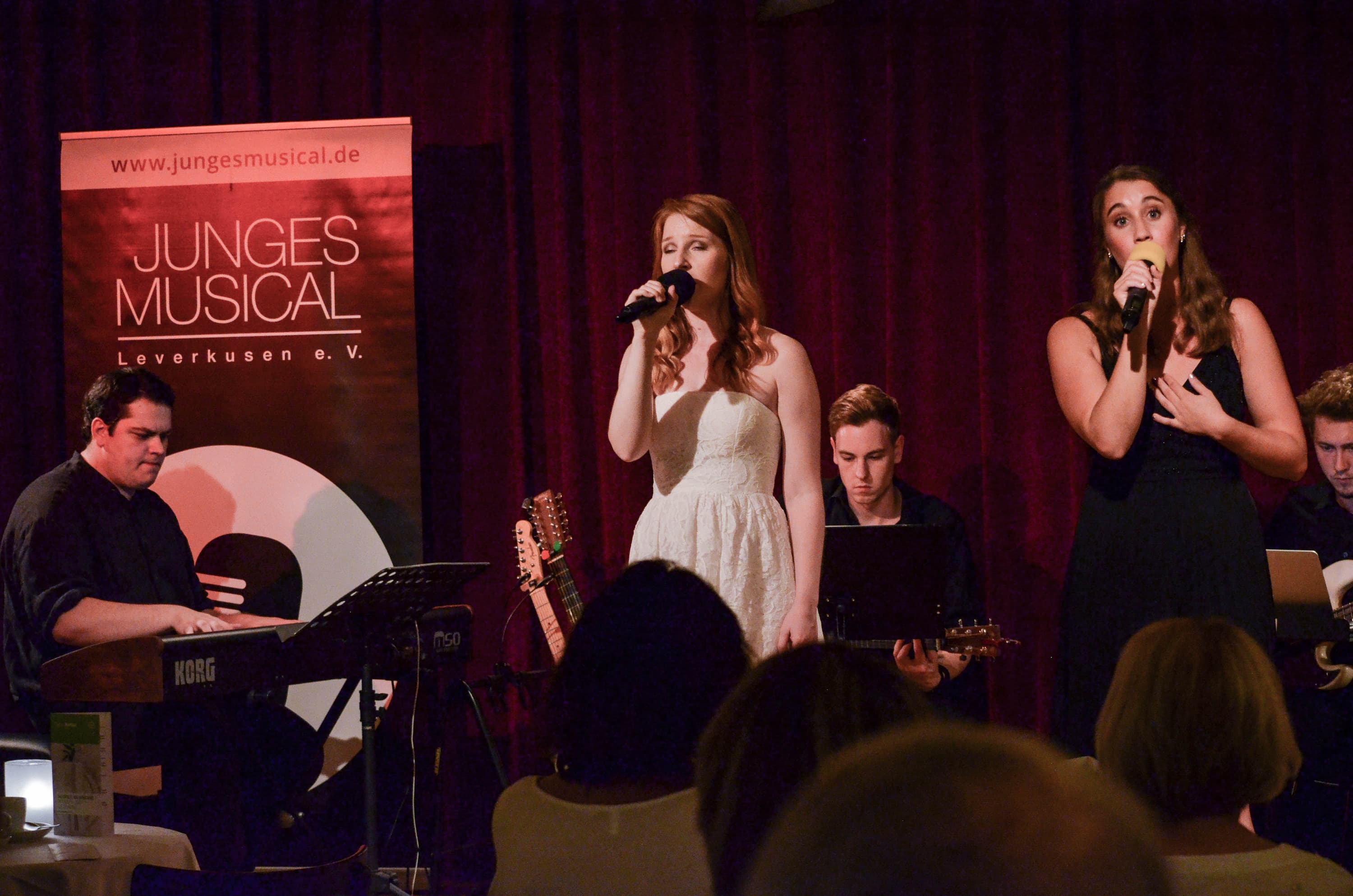 Zwei Sängerinnen stehen im Abendkleid auf einer kleinen Bühne mit einer Band im Hintergrund.