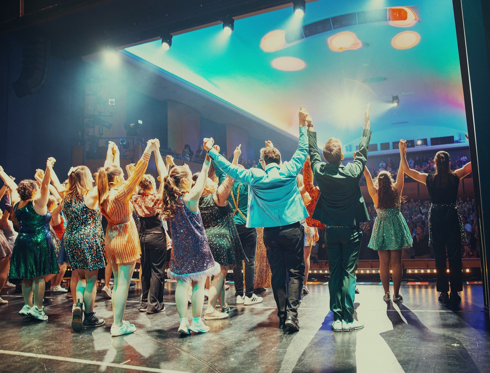 Gruppe von Darstellerinnen und Darstellern steht am Ende einer Bühnenaufführung nebeneinander, hält sich an den Händen und hebt die Arme, während das Publikum im Hintergrund applaudiert und bunte Scheinwerfer die Szene beleuchten.