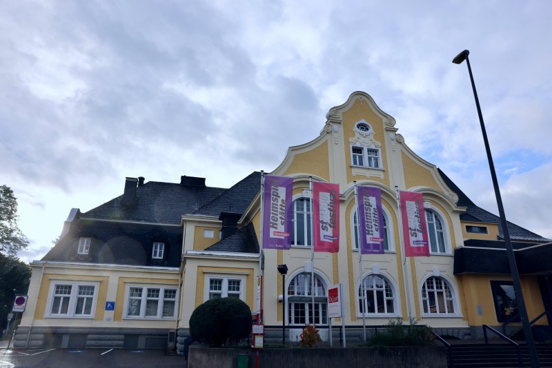 Historisches gelbes Theatergebäude mit geschwungenem Giebel und Fahnen vor bewölktem Himmel.
