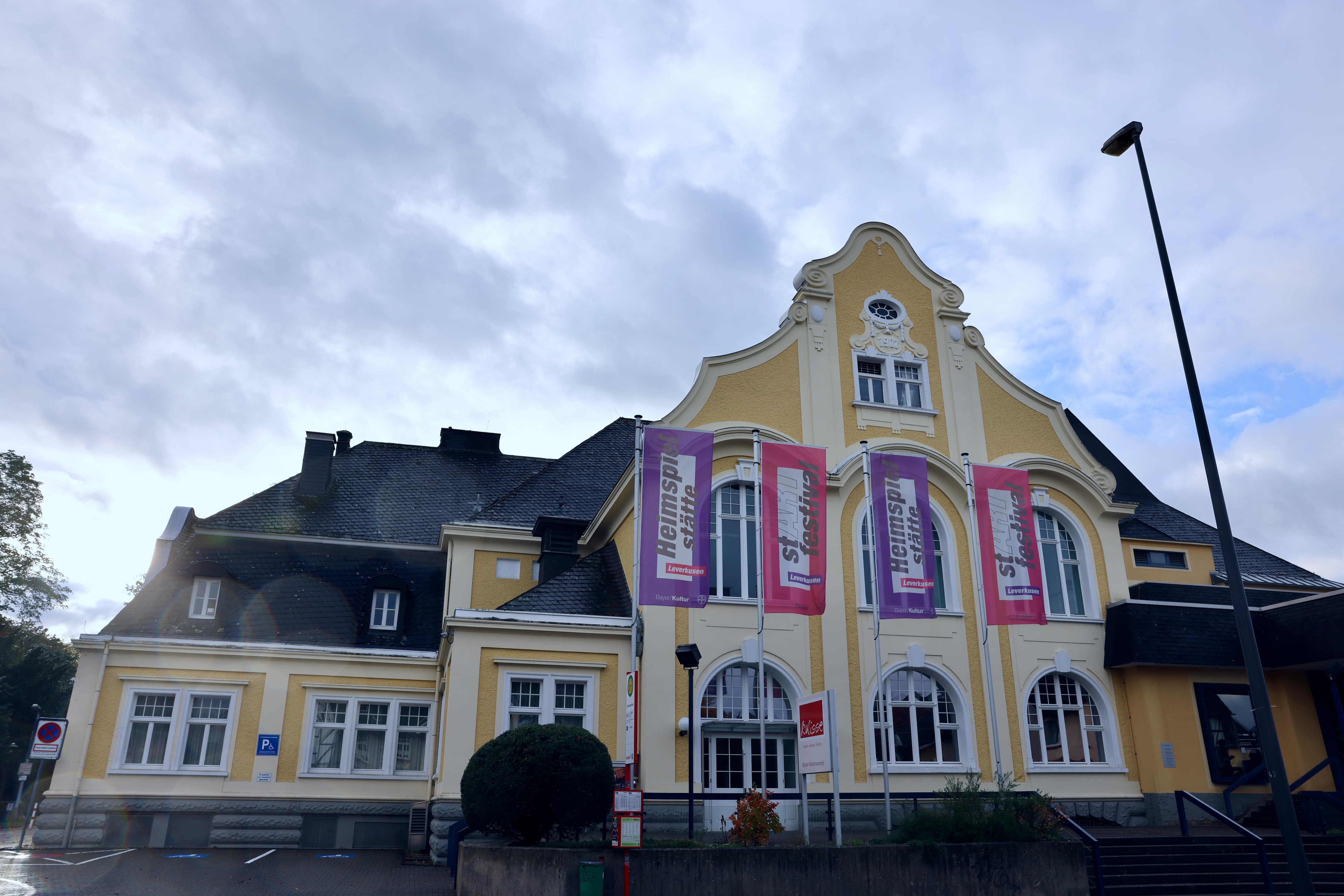 Historisches gelbes Theatergebäude mit geschwungenem Giebel und Fahnen vor bewölktem Himmel.