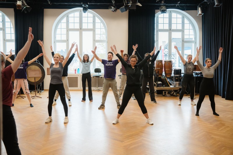 Gruppe von Erwachsenen beim Warmup in hellem Saal mit erhobenen Armen vor großen Fenstern.