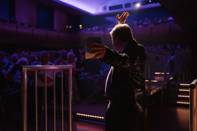 Blick von der Bühne in den Theatersaal: Darsteller im Scheinwerferlicht vor voll besetztem Publikum, stimmungsvolle Abendatmosphäre.