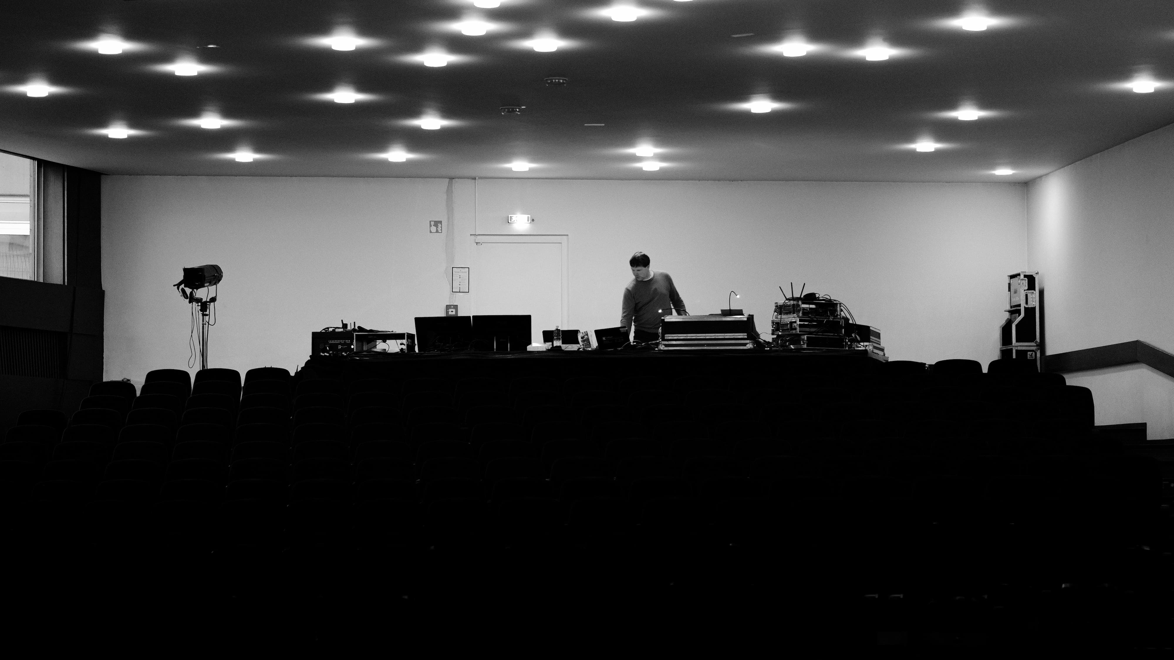 Leerer Theatersaal mit Blick auf den Technikbereich im Hintergrund. Eine Person steht zwischen Licht- und Tontechnik, vor ihr mehrere Monitore und Geräte zur Vorbereitung einer Aufführung.