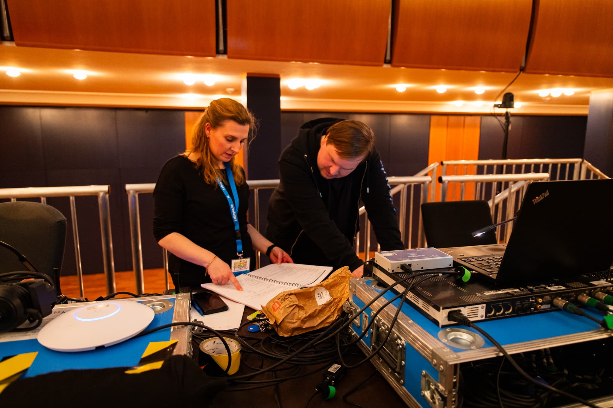 Zwei Personen im Technikbereich prüfen einen Ablaufplan. Vor ihnen stehen Laptop, Audiotechnik und Kabel auf einem Arbeitstisch im Theatersaal.