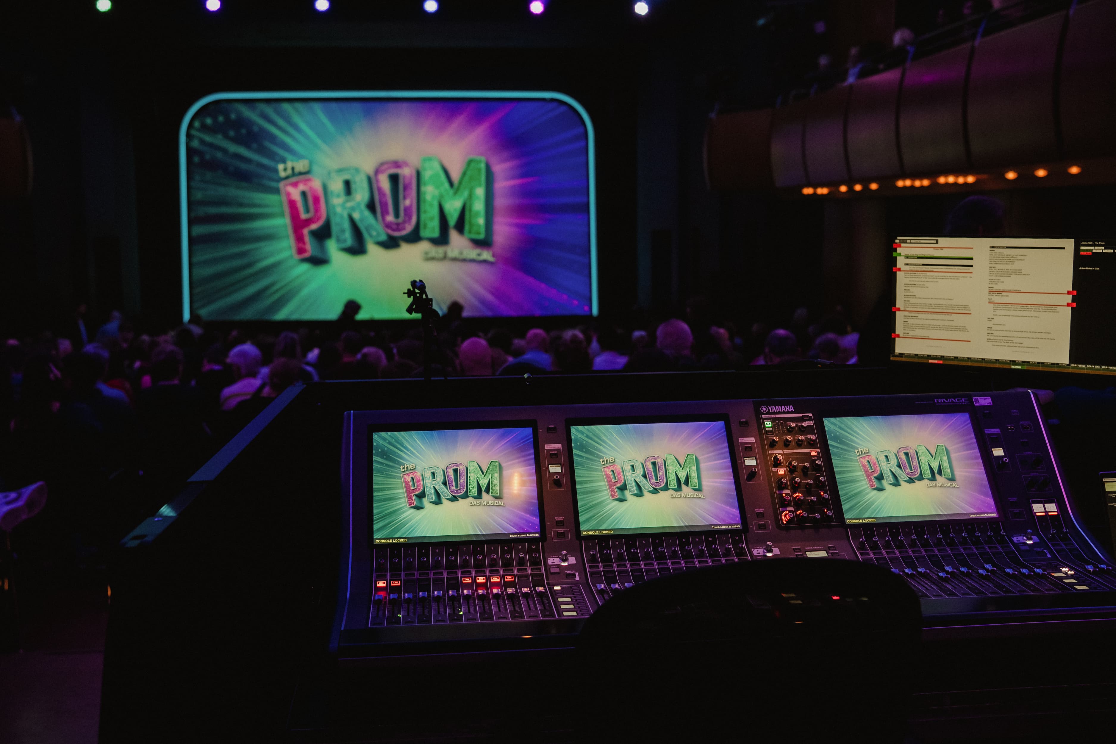 Blick aus der Technik auf eine Theaterbühne mit großer Leinwand, auf der "The Prom: Das Musical" angezeigt wird; im Vordergrund ein Mischpult mit mehreren Monitoren, Publikum im Hintergrund.