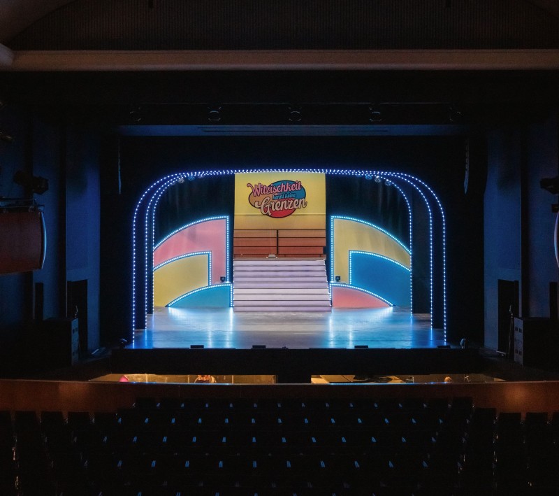 Leerer Theatersaal mit Blick auf eine beleuchtete Bühne. Buntes Bühnenbild mit Treppe und LED-Rahmen, darüber das Schild „Witzigkeit kennt keine Grenzen“.