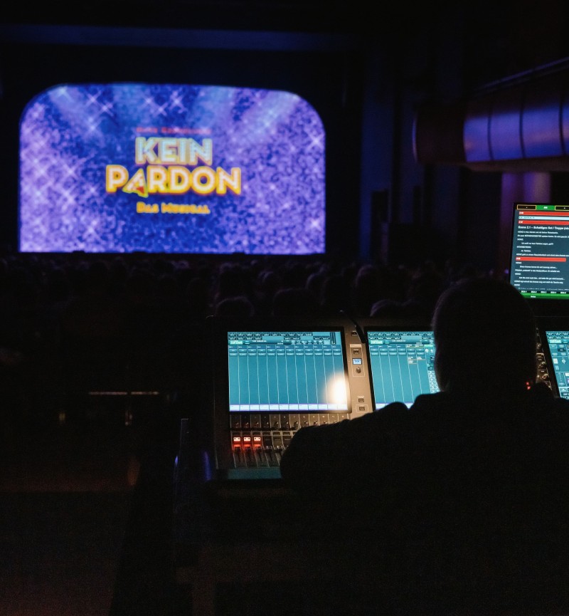 Blick aus dem Technikbereich in einen voll besetzten Theatersaal. Im Vordergrund stehen beleuchtete Licht- und Tonpulte, im Hintergrund ist auf einer großen Bühne mit Projektion zu sehen.