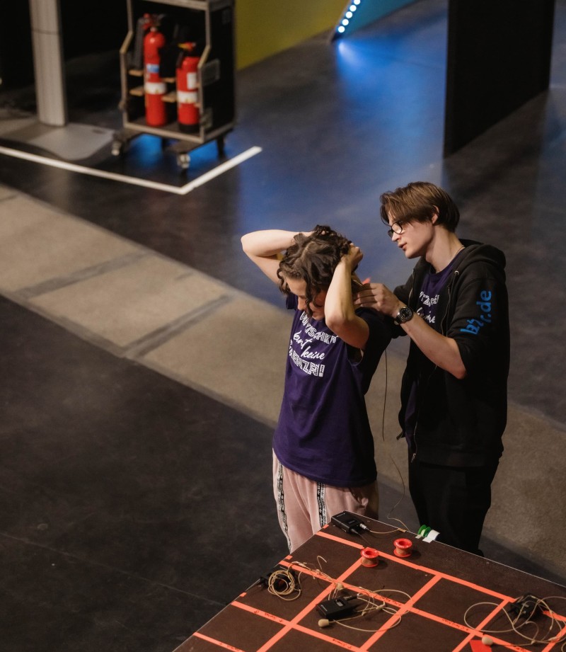 Zwei Personen bereiten hinter der Bühne ein Headset-Mikrofon vor. Auf einem Tisch liegen weitere Funkmikros und Zubehör für die Aufführung.