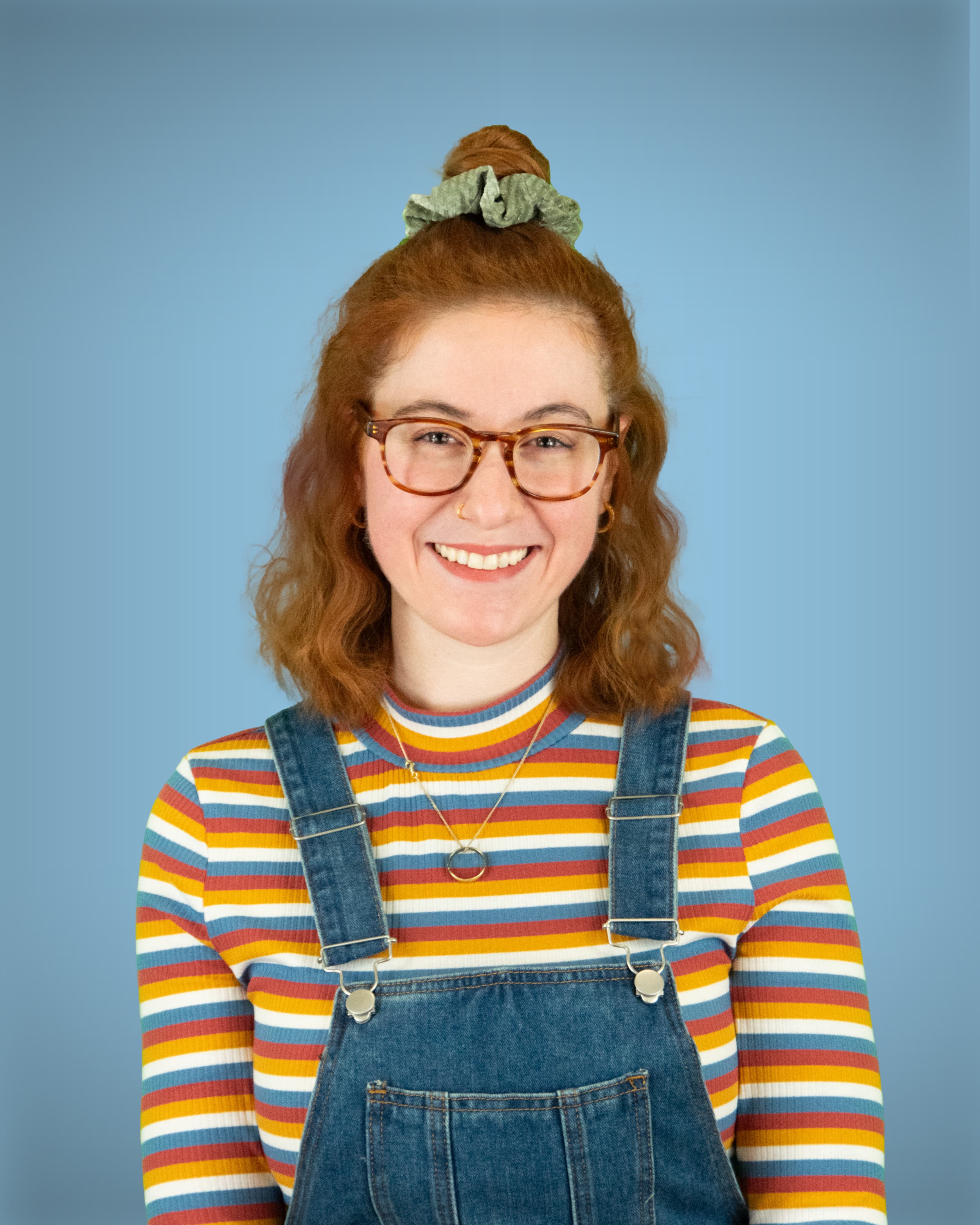 Eine junge Person mit rötlichen Haaren, Brille in Schildpatt-Optik und einem bunt gestreiften Pullover unter einer Jeanslatzhose blickt lächelnd in die Kamera.