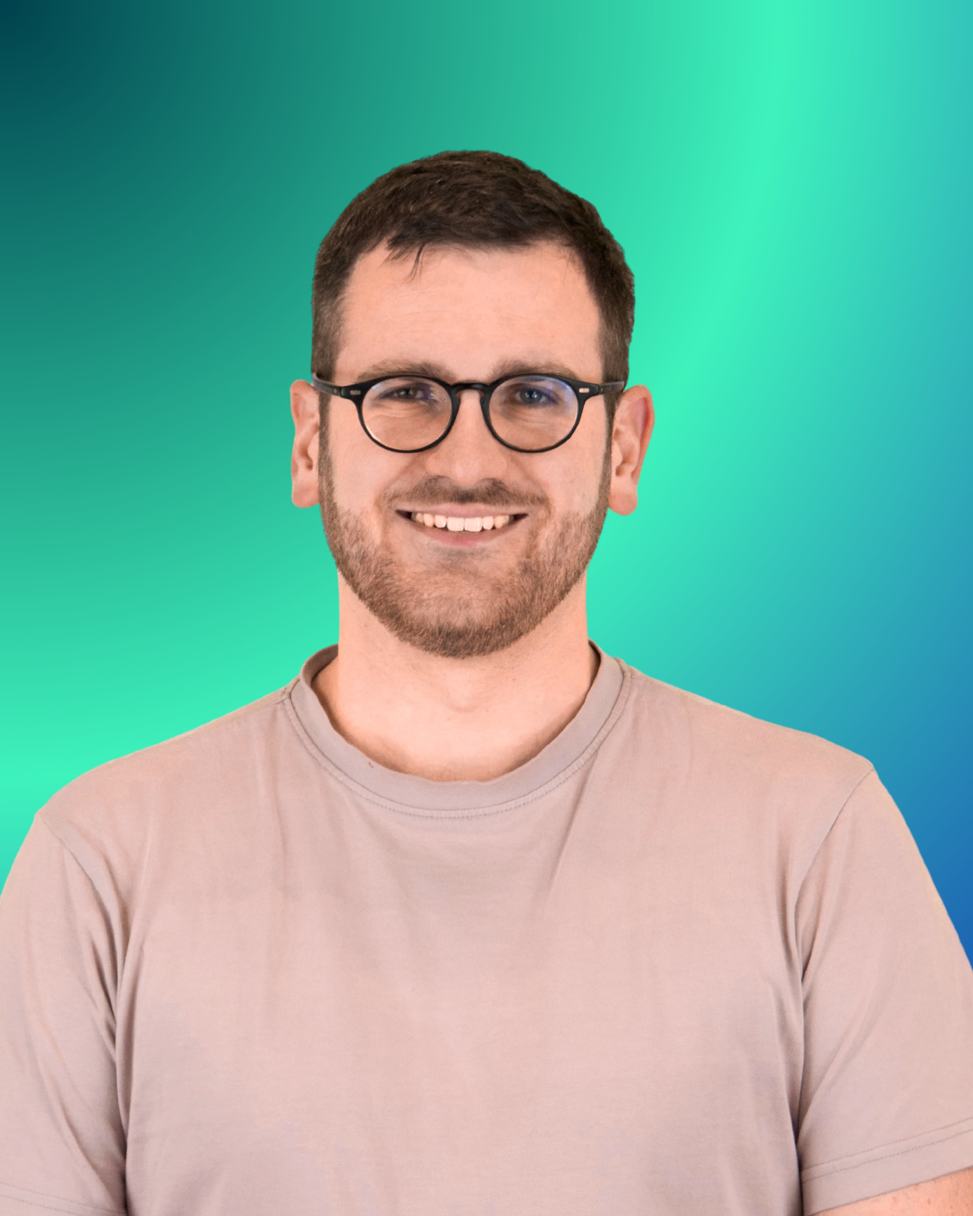Ein junger Mann mit kurzen dunklen Haaren, Brille und Dreitagebart blickt lächelnd in die Kamera; er trägt ein hellbeiges T-Shirt.
