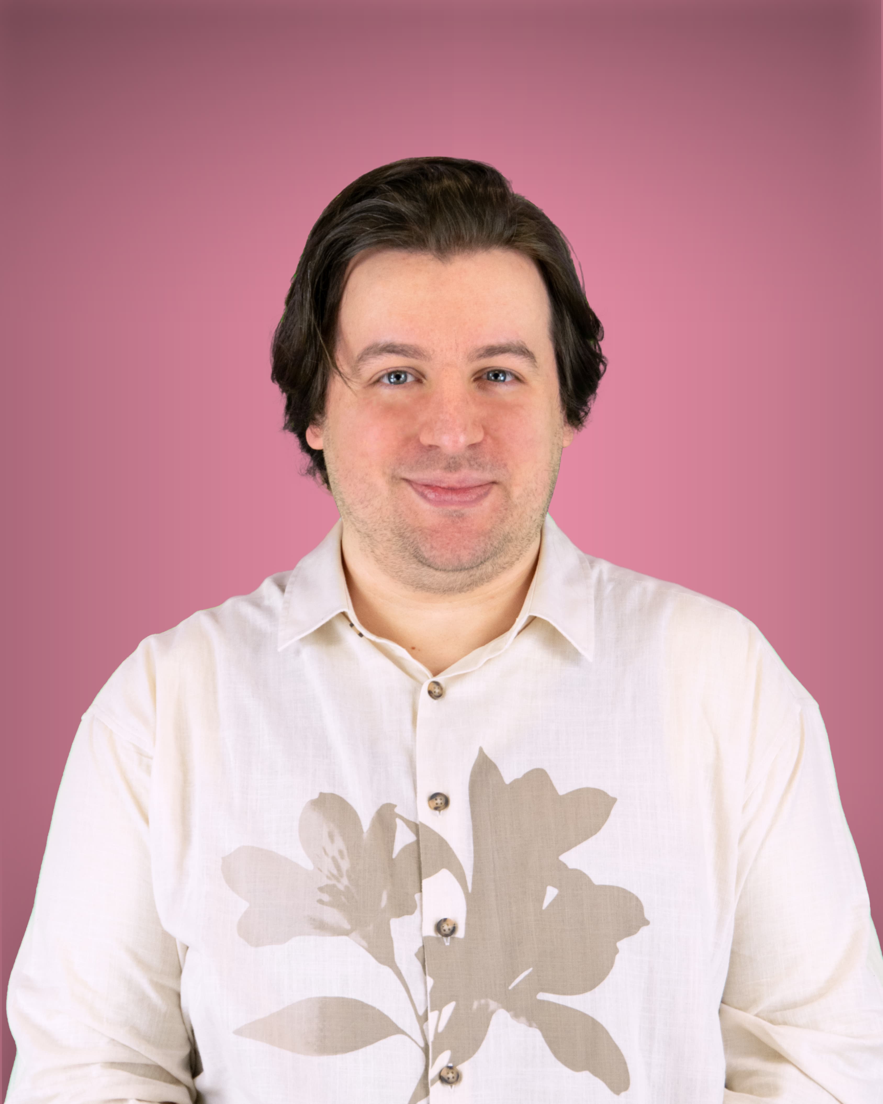 Ein Mann mit blauen Augen und dunklen Haaren blickt mit einem geschlossenen Lächeln in die Kamera; er trägt ein helles Hemd mit einem großen floralen Muster.
