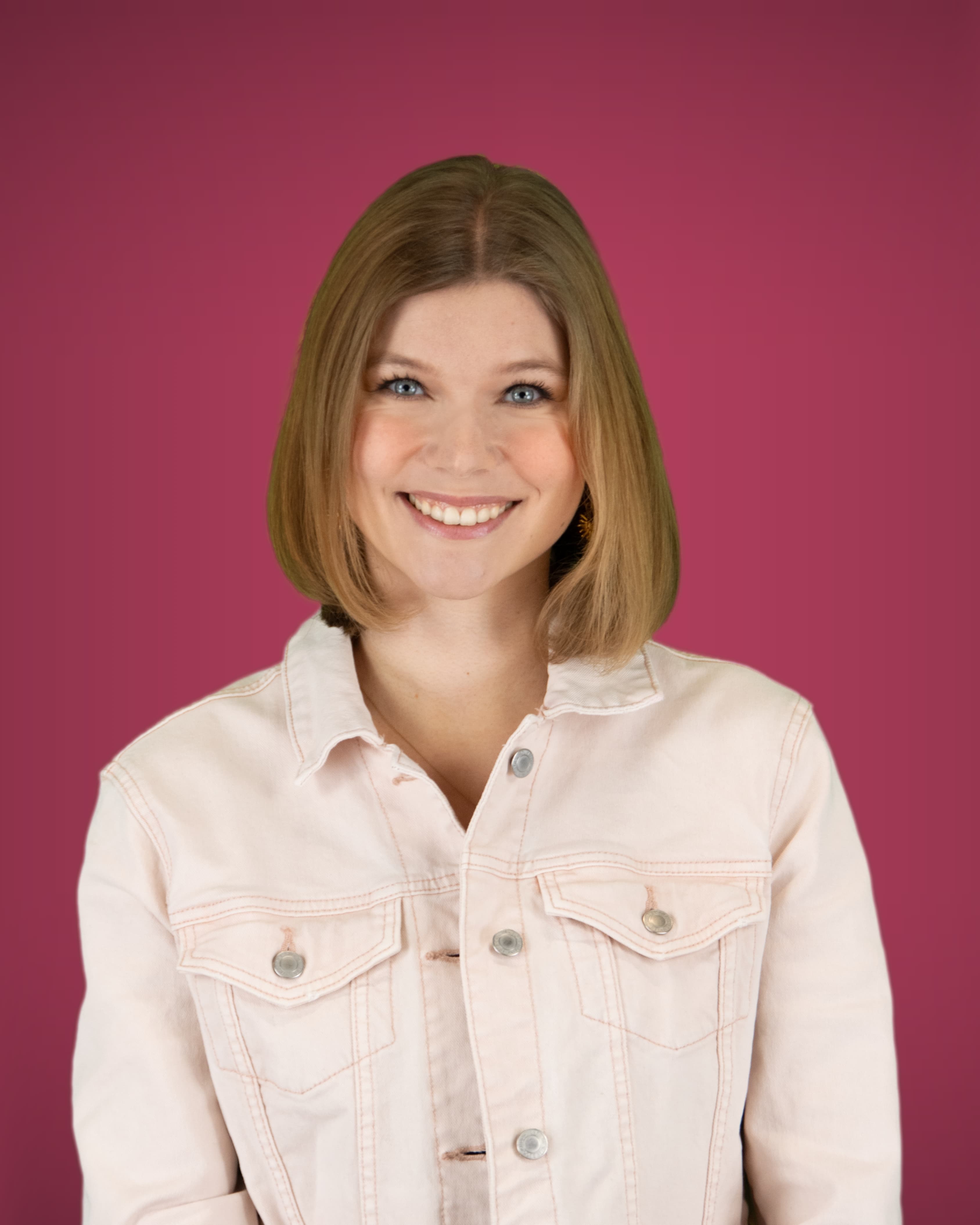 Eine junge Frau mit blauen Augen und einem hellbraunen Bob blickt lächelnd in die Kamera; sie trägt eine hellrosa Jeansjacke.