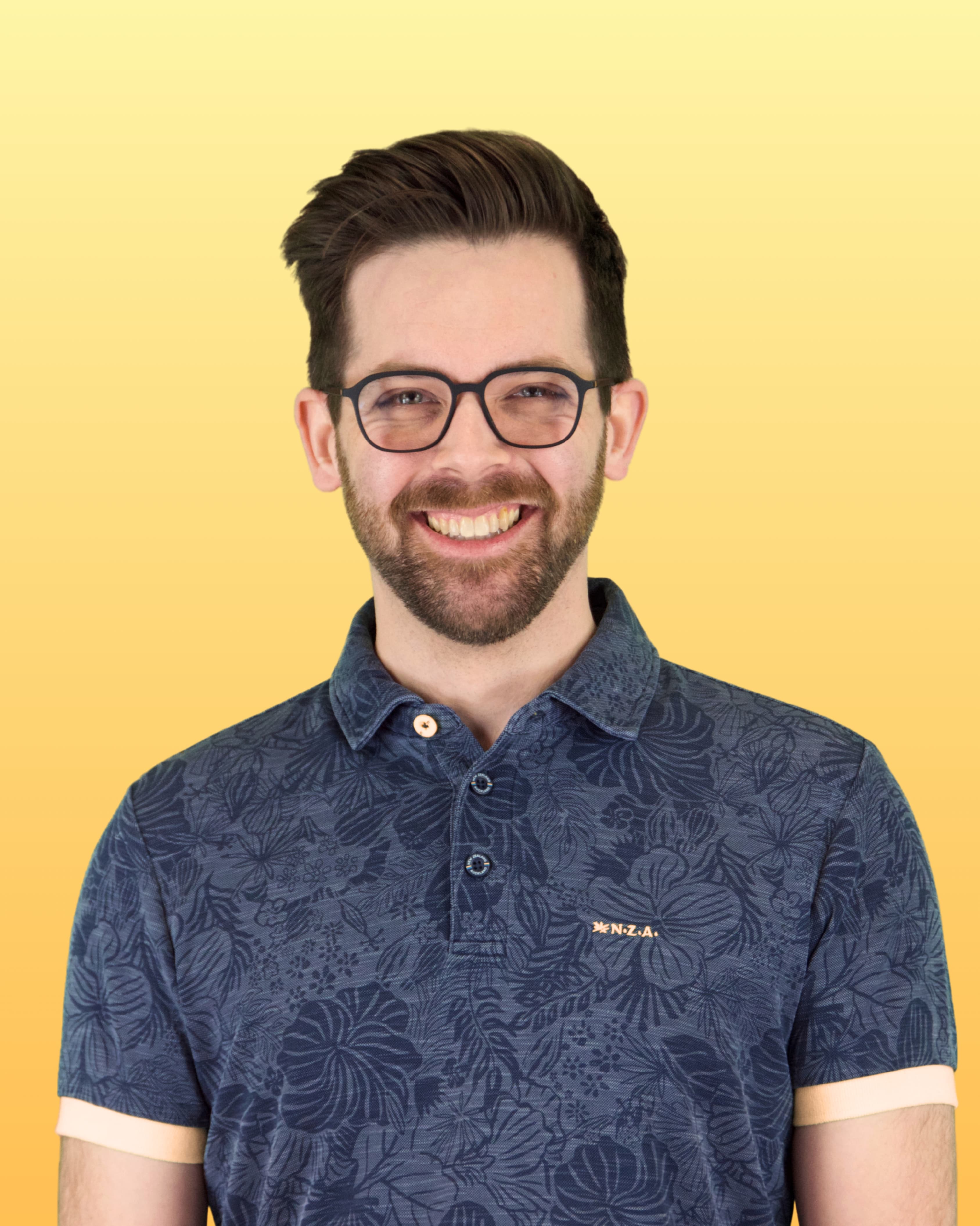 Ein junger Mann mit hochgesteilten dunklen Haaren, Brille und gepflegtem Bart blickt lachend in die Kamera; er trägt ein gemustertes Poloshirt.
