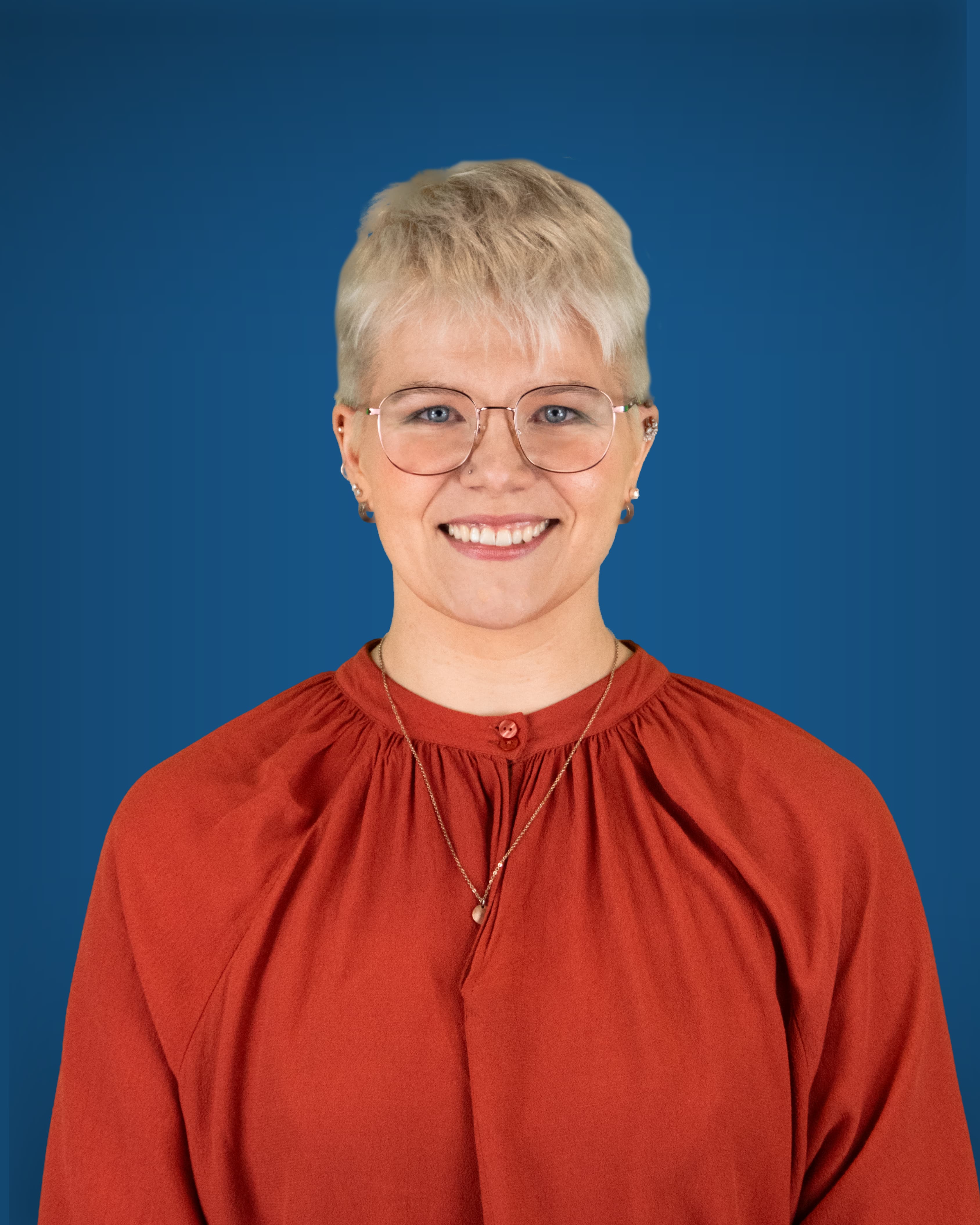 Eine junge Person mit platinblondem Kurzhaarschnitt, blauen Augen und runder Brille blickt lächelnd in die Kamera; sie trägt eine rostrote Bluse.