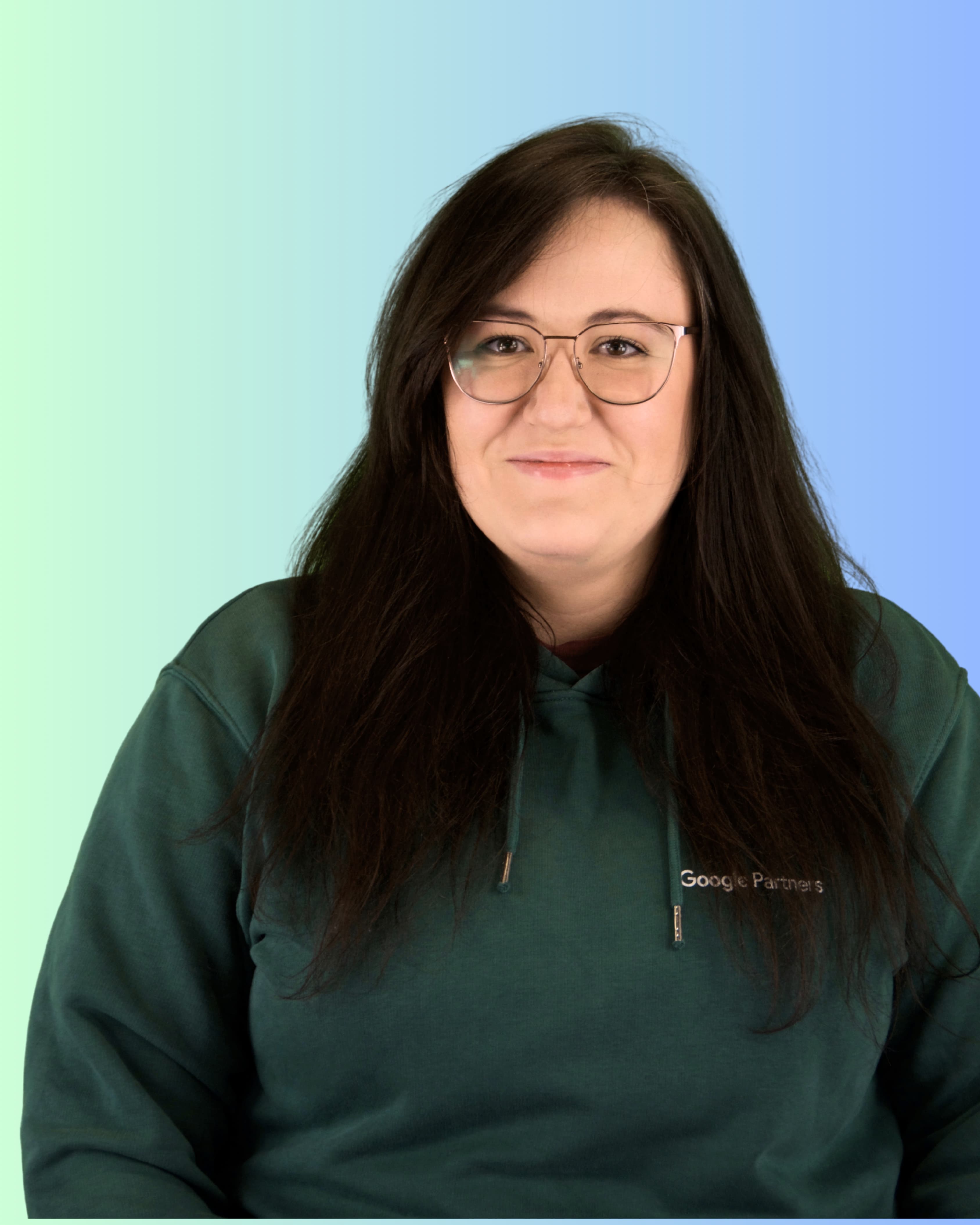 Eine Person blickt mit Brille und langen, dunklen Haaren direkt in die Kamera und trägt einen dunkelgrünen Hoodie mit dem sichtbaren Schriftzug Google Partners.