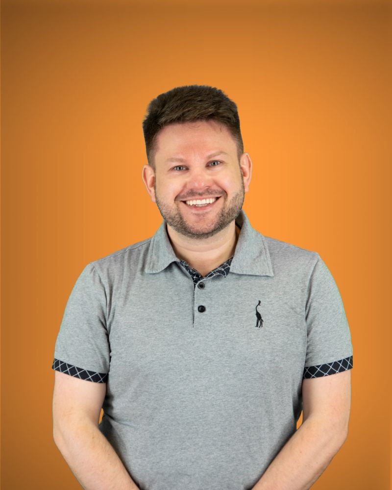 Ein Mann blickt lächelnd direkt in die Kamera; er trägt ein graues Poloshirt mit einem kleinen Giraffen-Logo auf der Brust sowie gemusterten Ärmelbündchen und Krageninnenseiten vor orangefarbenem Hintergrund.