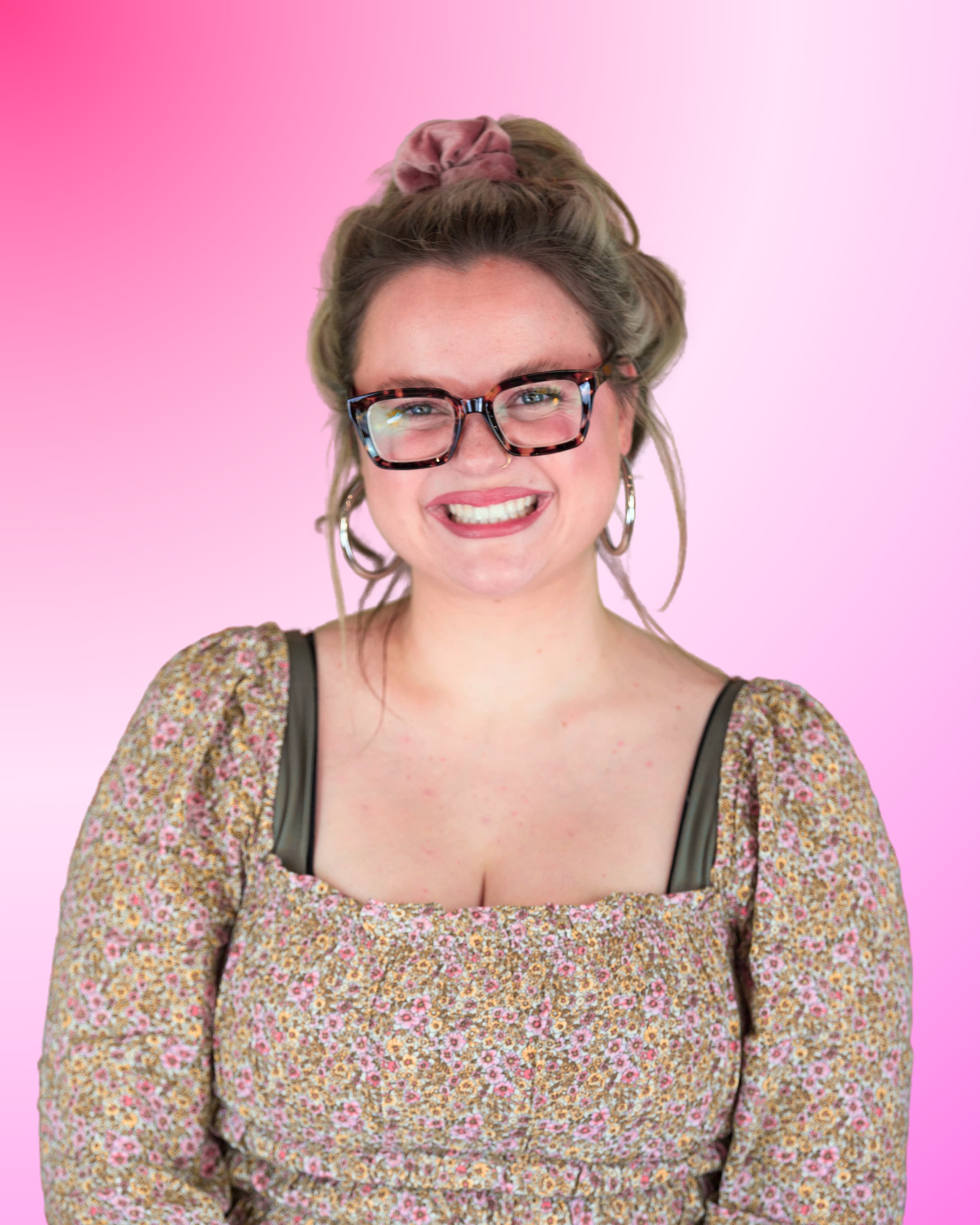 Eine Person blickt lächelnd in die Kamera. Sie trägt eine eckige Brille mit Schildpatt-Muster, große Kreolen-Ohrringe und ein florales, feingemustertes Oberteil. Ihre Haare sind mit einem hellen Haargummi hochgesteckt.