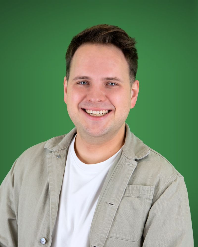 Eine Person mit blauen Augen und kurzen, hochgestellten braunen Haaren blickt direkt und lächelnd in die Kamera. Sie trägt ein weißes T-Shirt unter einer offenen, hellgrauen Jacke aus festem Stoff.