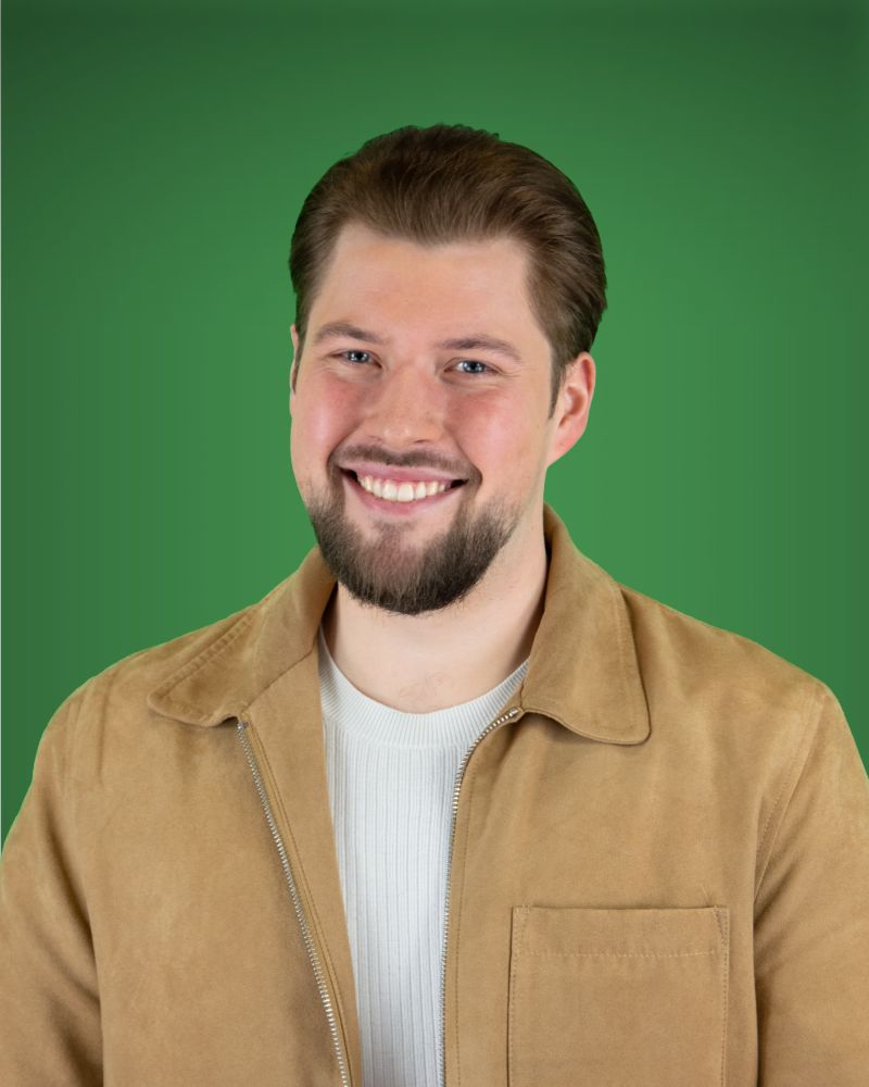 Eine Person mit blauen Augen und einem gestutzten Vollbart blickt lächelnd in die Kamera. Sie trägt ein weißes, fein geripptes Oberteil unter einer leicht geöffneten, beigen Jacke mit Reißverschluss und Kragen.