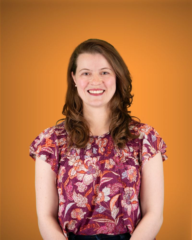 Eine Person mit leicht gewellten, braunen Haaren blickt lächelnd in die Kamera. Sie trägt eine kurzärmelige Bluse mit floralem Muster in Lila-, Orange- und Weißtönen sowie kleine Perlenohrstecker.