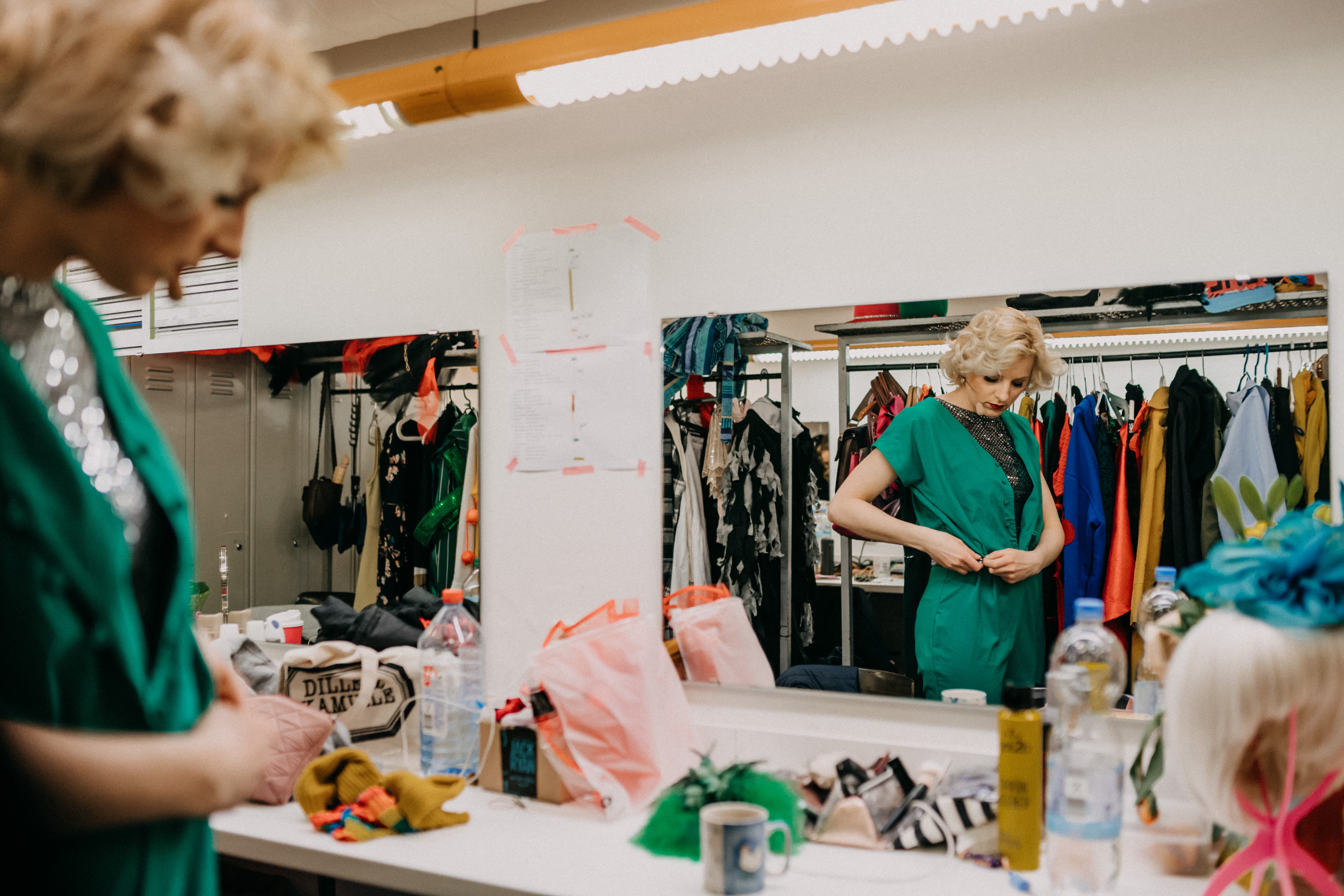 Eine Frau steht vor dem Spiegel und zeiht sich ein Kostüm an. Im Hintergrund hängen mehrere Kleider an Gardinenstangen.