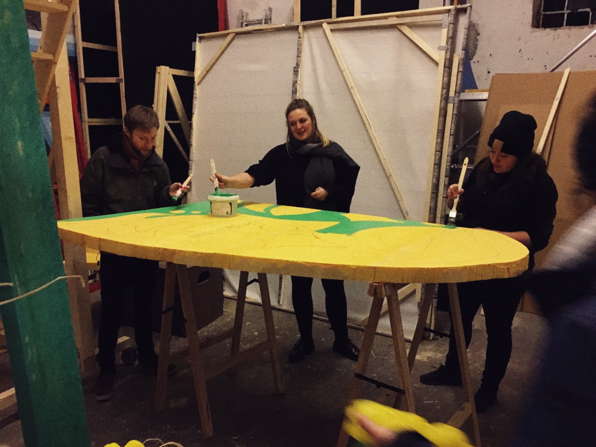 Drei Menschen stehen in einer Halle und bemalen ein Bühnenbildelement.