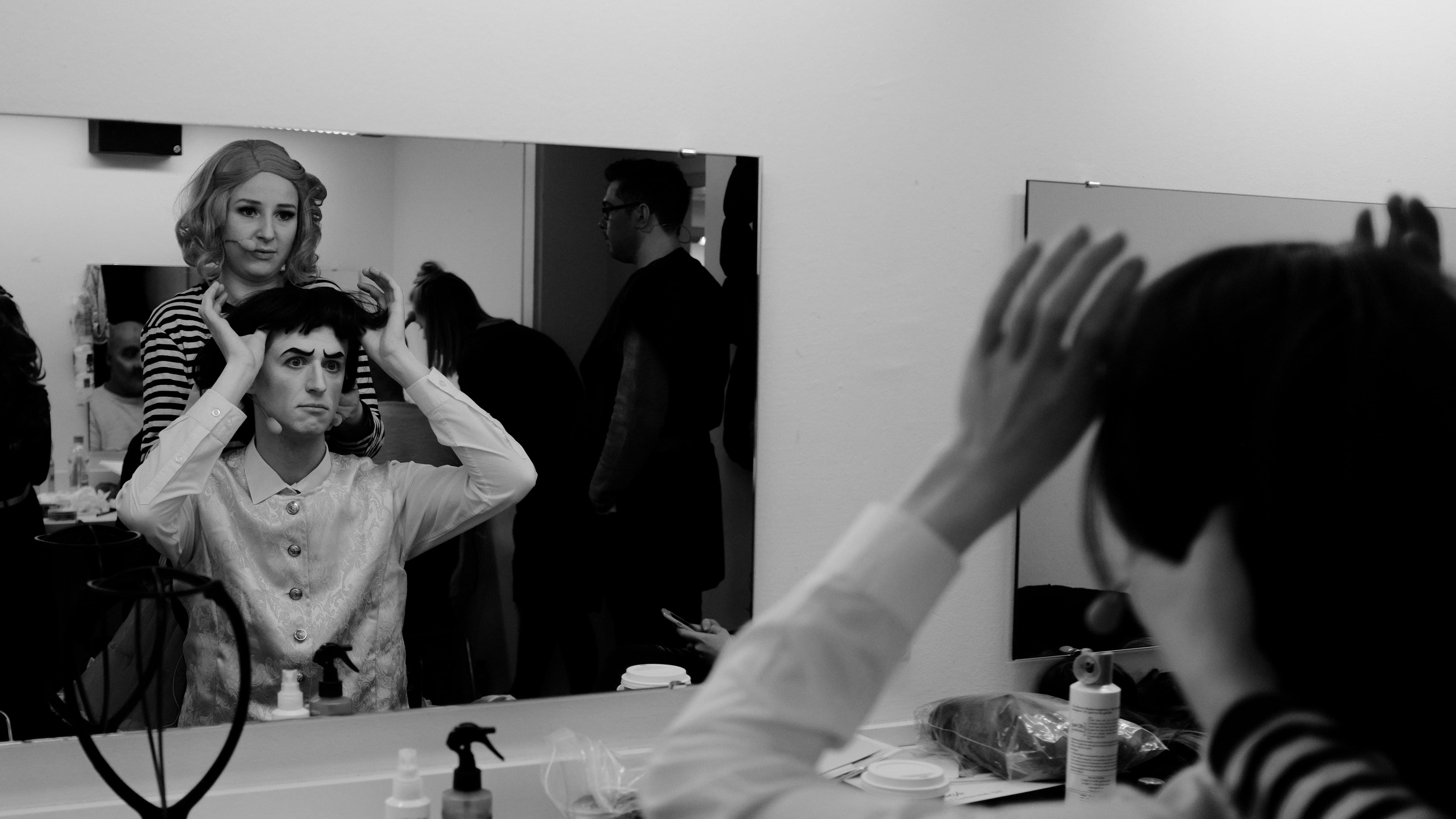 Eine Person zieht einer sitzenden Person eine Perücke über den Kopf.