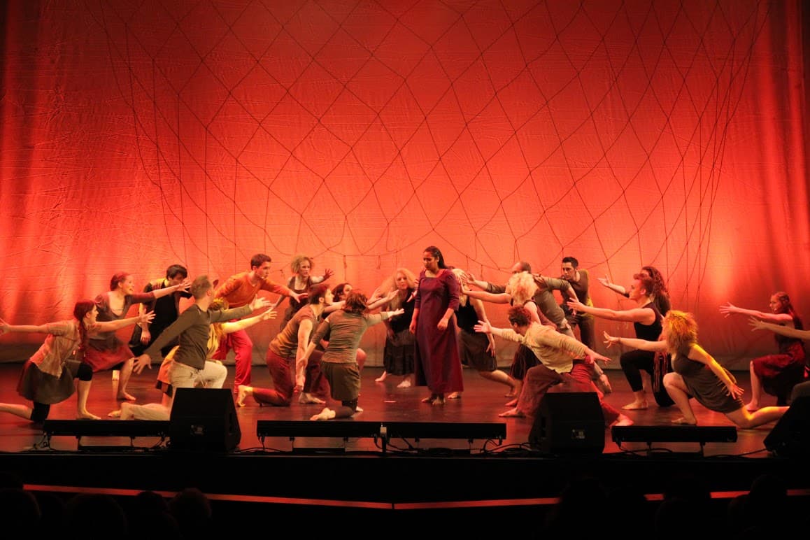 Eine Darstellerin wird von dem restlichen Ensemble in Landkleidung auf der Bühne flehend umzingelnd.