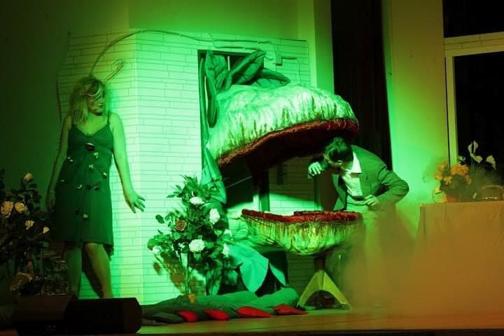 Ein Darsteller schaut in das weit geöffnete Maul einer riesigen fleischfressenden Pflanze. Neben der Pflanze steht eine Frau in einem grünen Kleid.