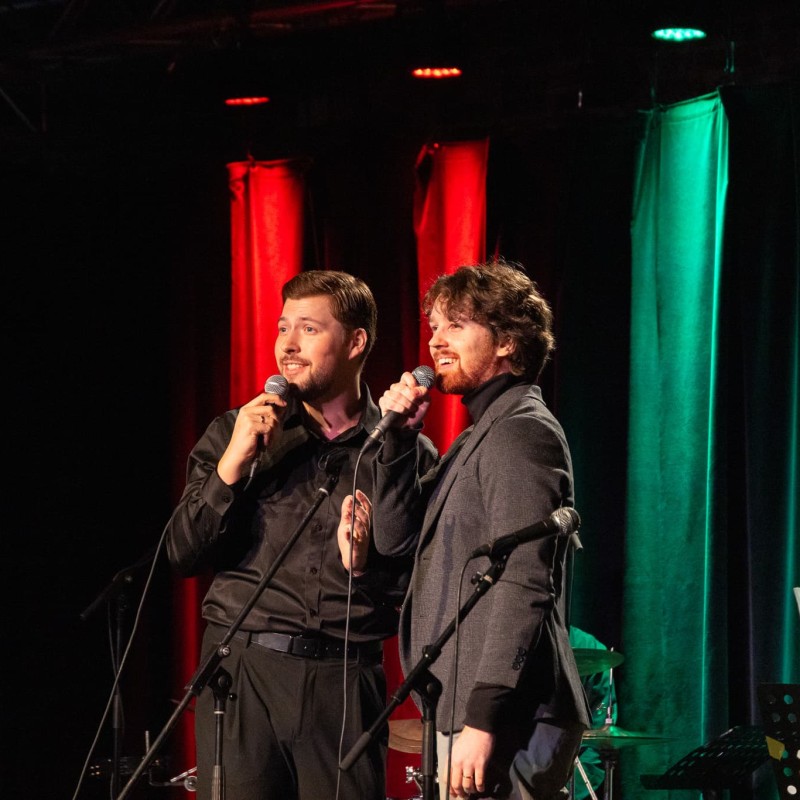 Zwei Männer singen gemeinsam auf einer Bühne mit Mikrofonen in der Hand, im Hintergrund farbig beleuchtete Vorhänge in Rot und Grün sowie ein Musiker am Schlagzeug.