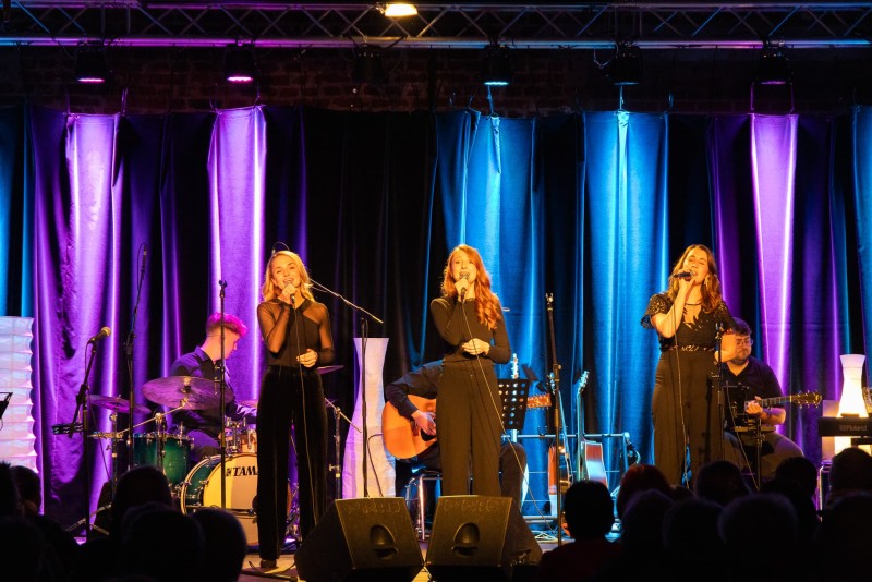 Drei Sängerinnen stehen nebeneinander auf einer Bühne und singen ins Mikrofon, begleitet von einer Live-Band mit Schlagzeug, Gitarre und Keyboard. Die Bühne ist in blau-violettes Licht getaucht, das Publikum ist im Vordergrund silhouettenhaft zu erkennen.