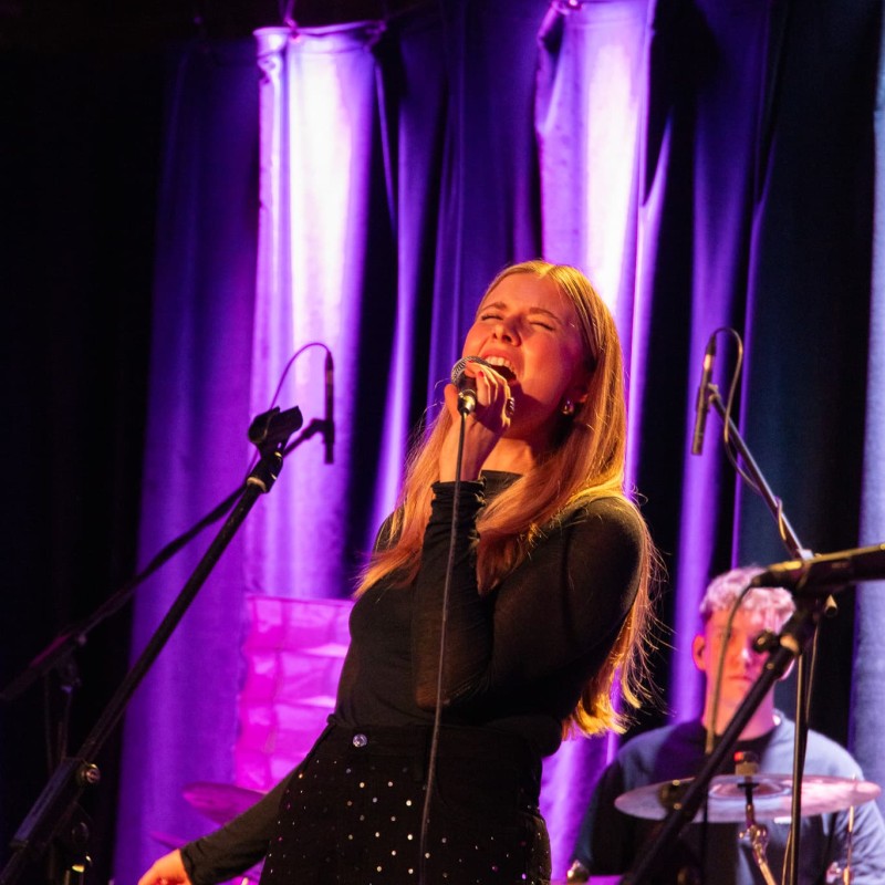 Junge Frau singt ausdrucksstark auf einer Bühne in violettem Licht, mit geschlossenen Augen und Mikrofon in der Hand. Im Hintergrund spielt ein Schlagzeuger, mehrere Mikrofone und Bühnenvorhänge sind sichtbar.