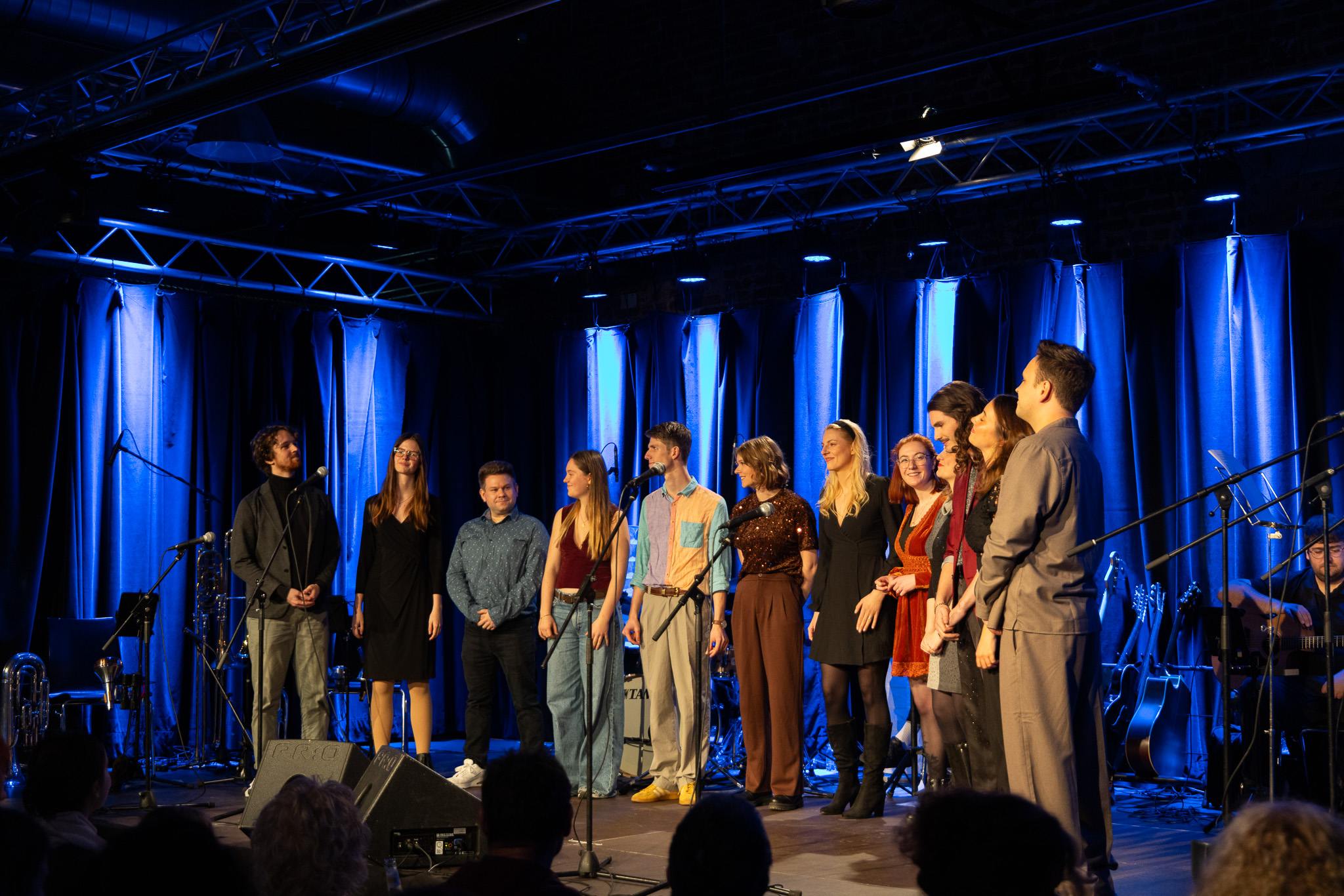 Zwölf Personen stehen in einer Reihe auf einer Bühne mit blau ausgeleuchtetem Vorhang. Die Gruppe trägt eine Mischung aus legerer und festlicher Kleidung, darunter Kleider, Hemden und Hosenanzüge. Rechts im Hintergrund sitzen Musiker zwischen Mikrofonen, Gitarren und einem E-Piano.