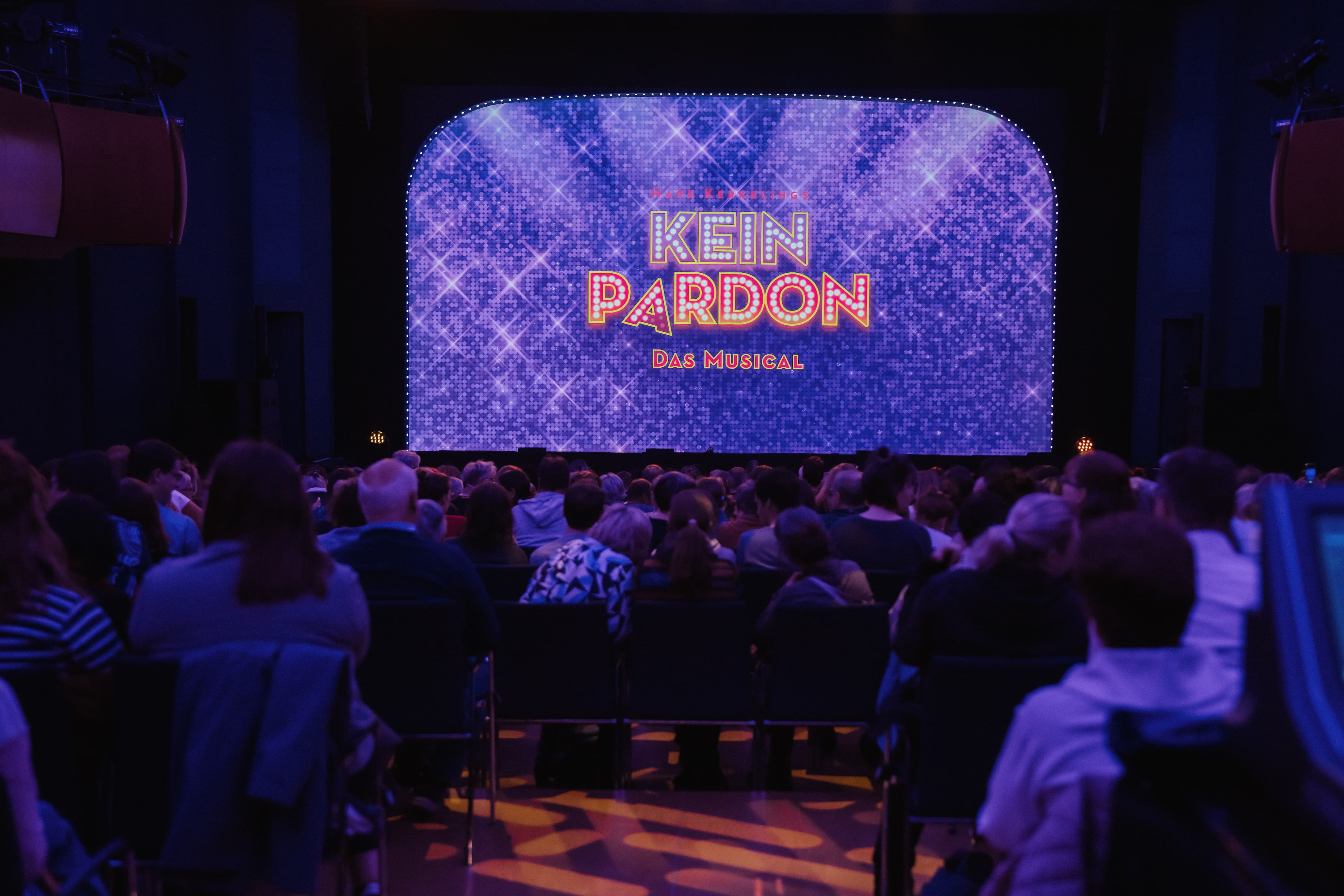 Publikum im Theatersaal vor einem glitzernden Vorhang mit der leuchtenden Aufschrift „Kein Pardon – Das Musical“, kurz vor Vorstellungsbeginn.