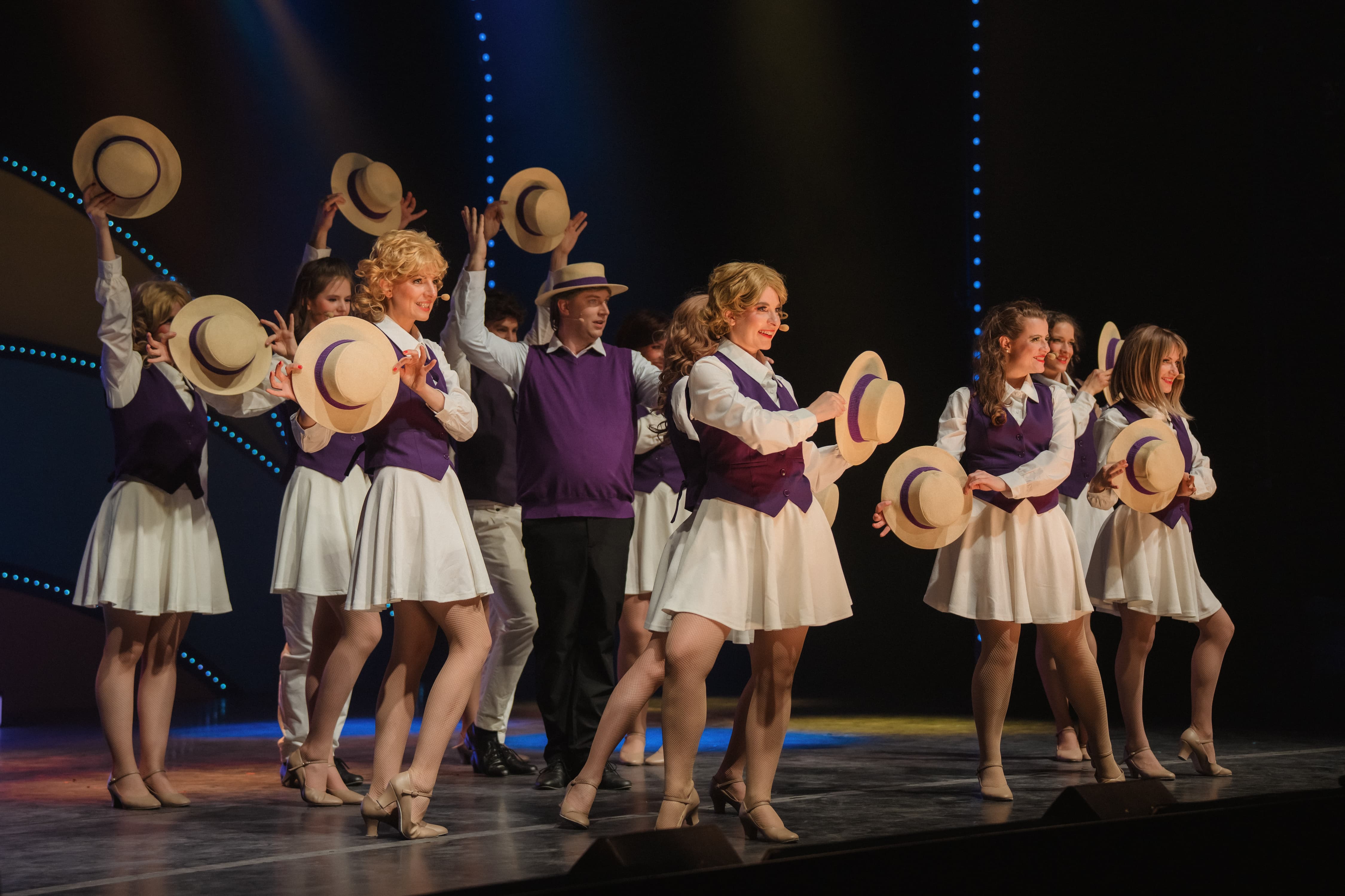 Tanzensemble in weißen Outfits mit lila Westen und Strohhüten auf einer Bühne, alle in synchroner Pose mit gehobenen oder präsentierten Hüten.