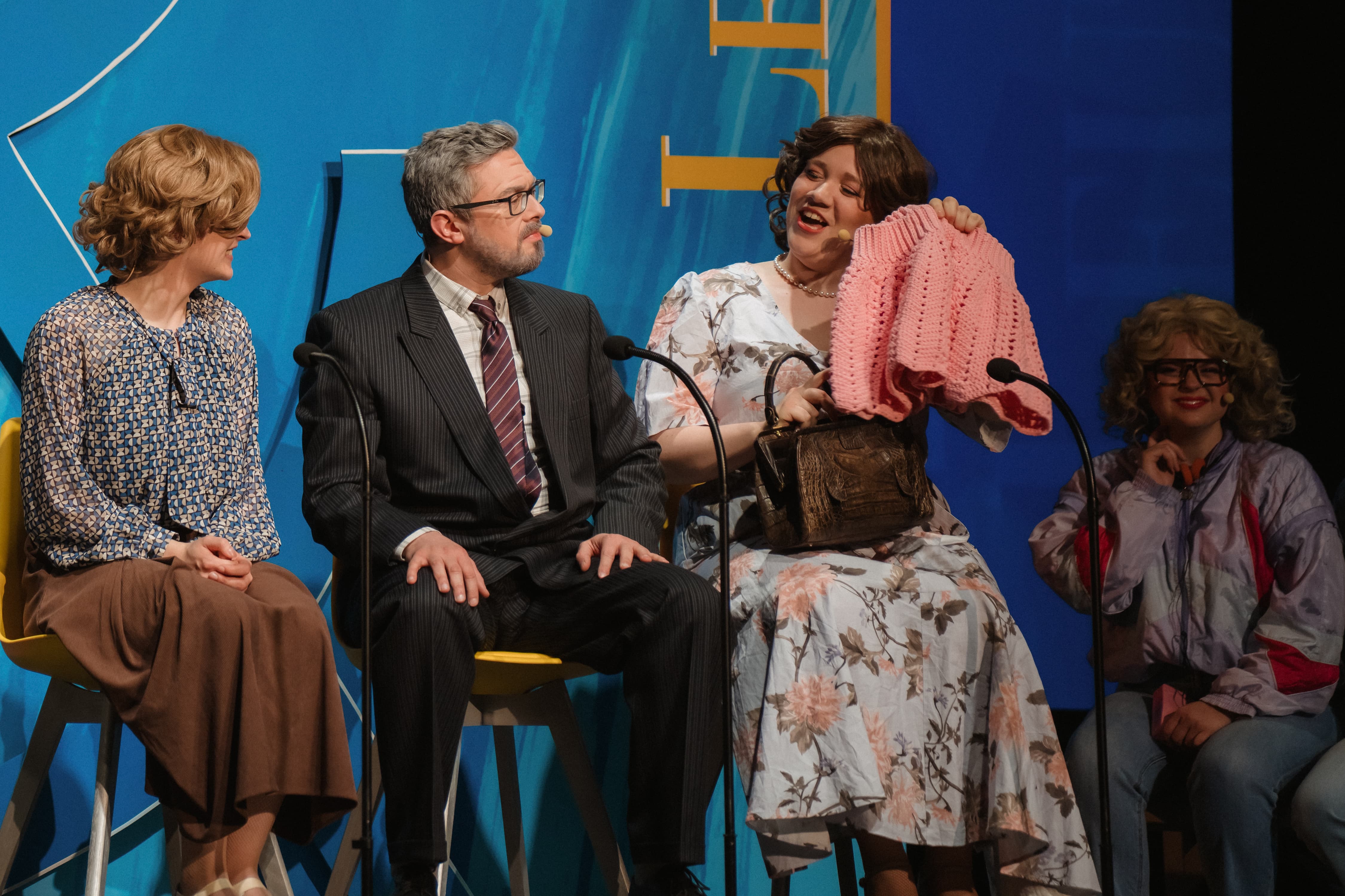 Drei Personen auf einer Bühne in Talkshow-Szenerie, eine Person zeigt lachend ein rosa Strickstück, alle in Alltags- oder Retro-Kleidung.