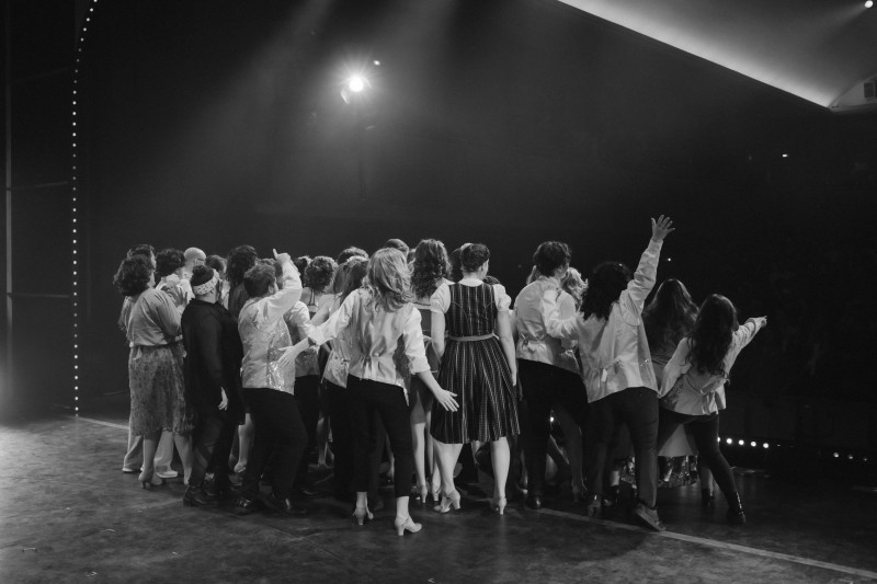 Gruppe von Menschen auf einer Bühne, jubelnd und mit dem Rücken zur Kamera, festlich beleuchtet, Publikum im Dunkeln.