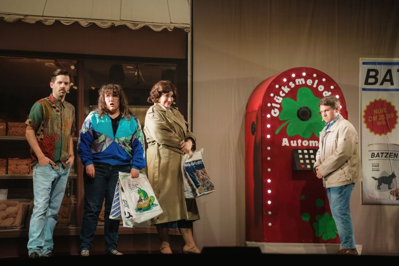 Vier Personen auf einer Bühne vor einem Geschäft und einem roten „Glücksmelodien-Automat“, alle tragen Einkaufstüten und blicken ernst oder überrascht.