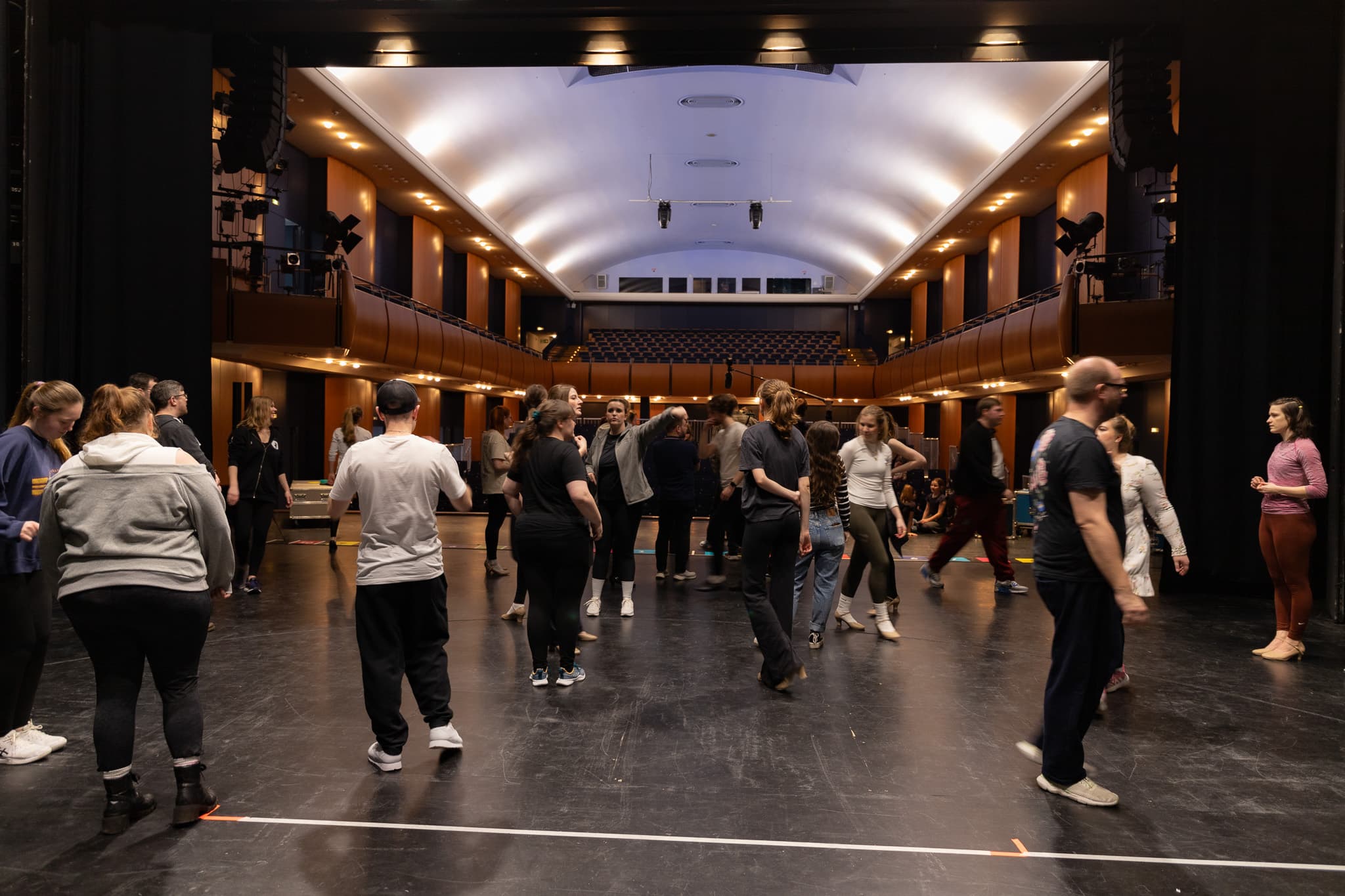 Eine Gruppe von Personen steht auf einer Theaterbühne vor einem beleuchteten Zuschauerraum und unterhält sich.