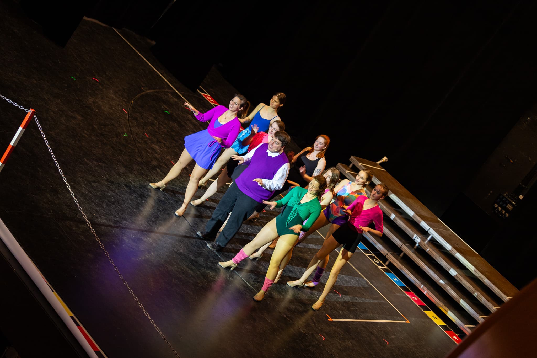 Tanzensemble auf einer Theaterbühne in farbenfrohen Kostümen, mitten in einer Choreografie. Im Hintergrund Bühnenpodeste und schwarze Kulissen.