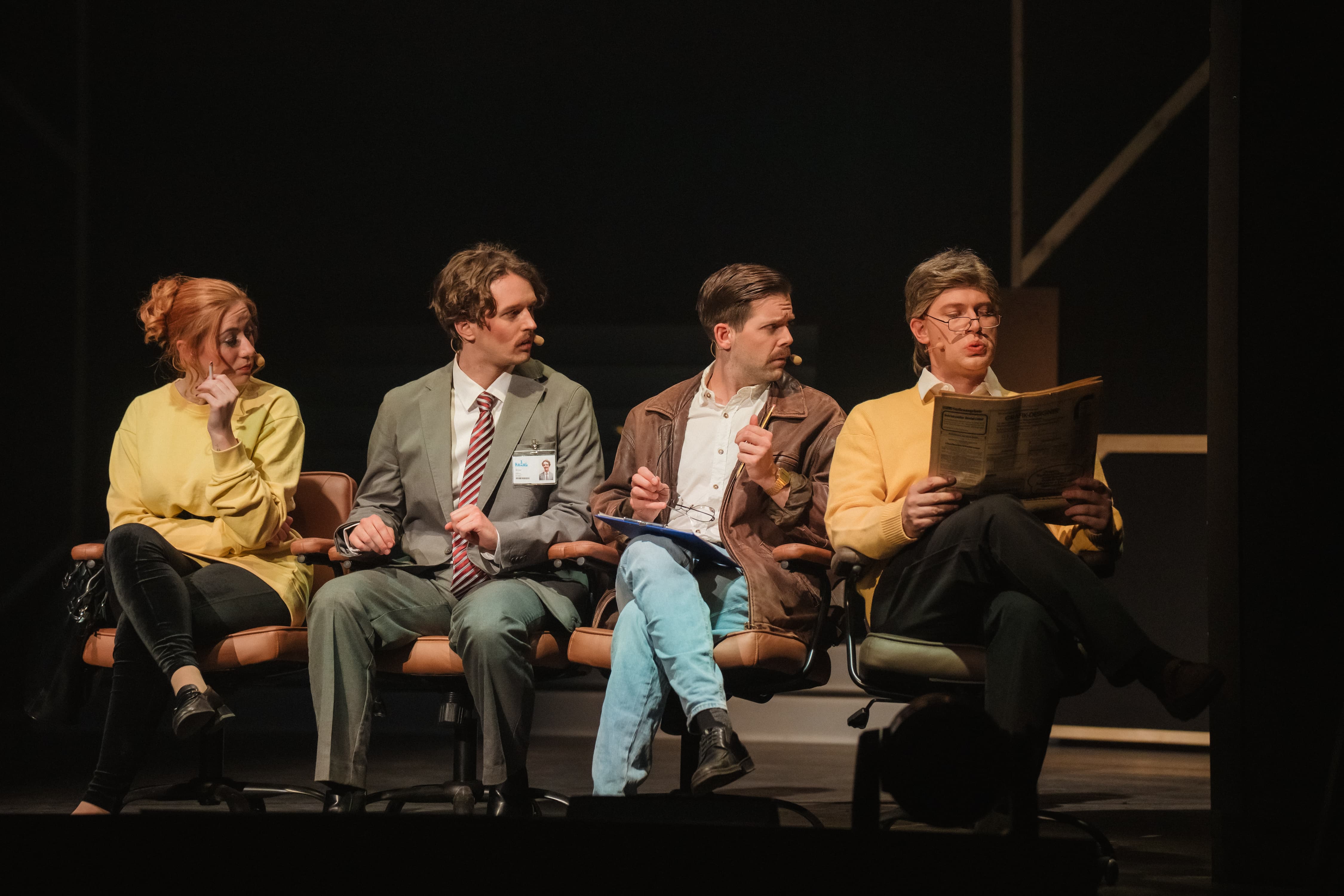 Vier Personen sitzen nebeneinander auf Bürostühlen, drei schauen gespannt zur Seite, eine liest gelassen Zeitung; alle tragen Headsets und Bühnenkostüme.