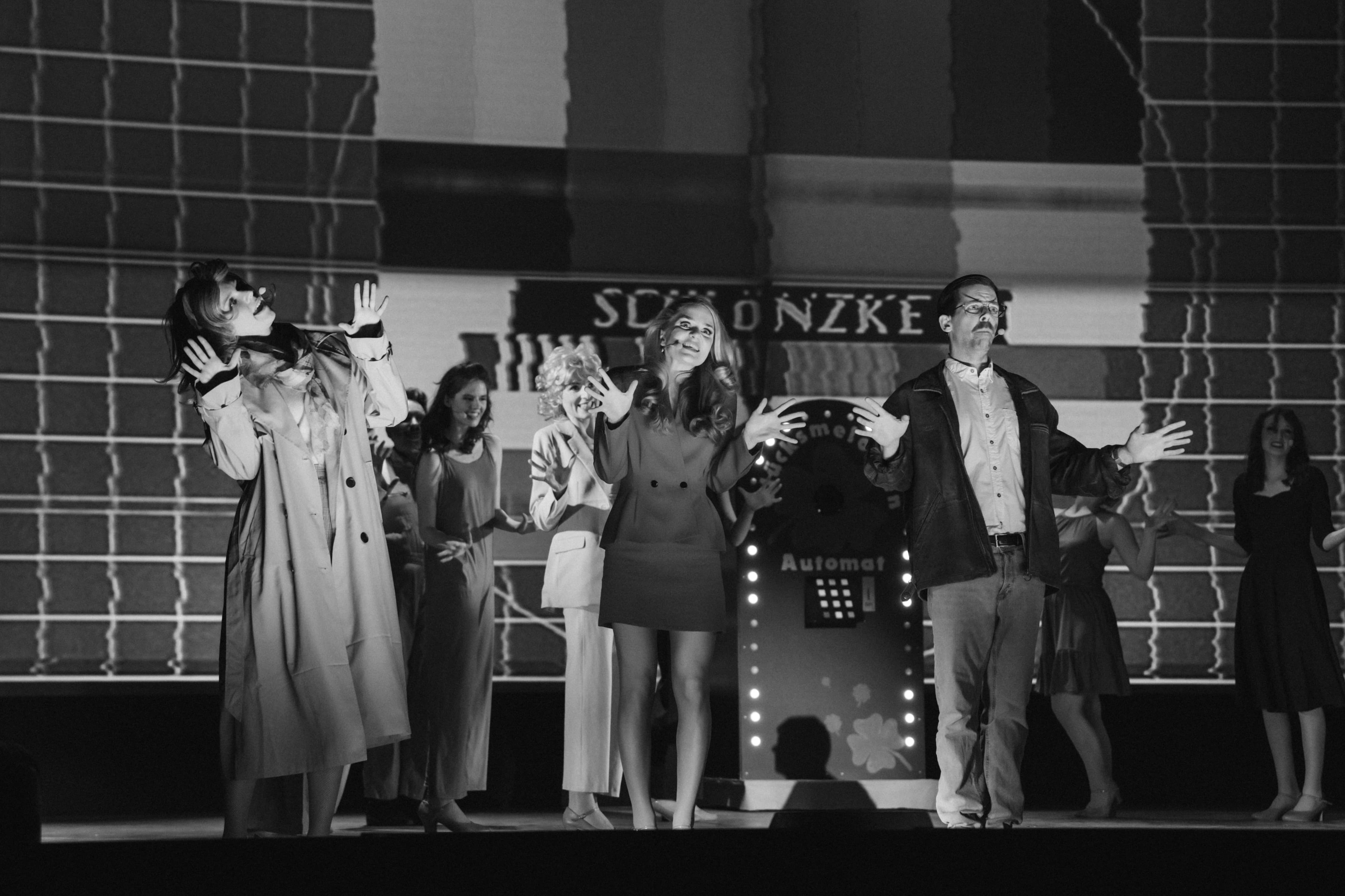 Mehrere Darstellende auf einer Bühne in szenischer Pose mit erhobenen Händen, im Hintergrund ein Testbild-Muster und ein Requisitenautomat.