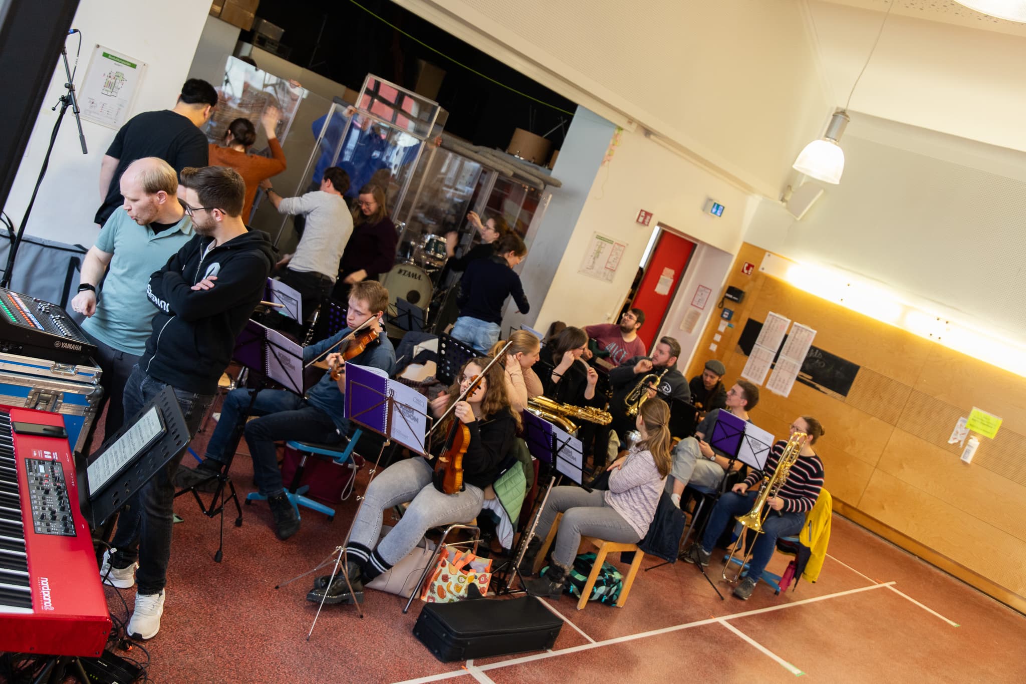 Eine Gruppe von Musikern probt in einem Raum mit verschiedenen Instrumenten, darunter Saxophone und Trompeten. Andere Personen bauen im Hintergrund die Ausrüstung auf.