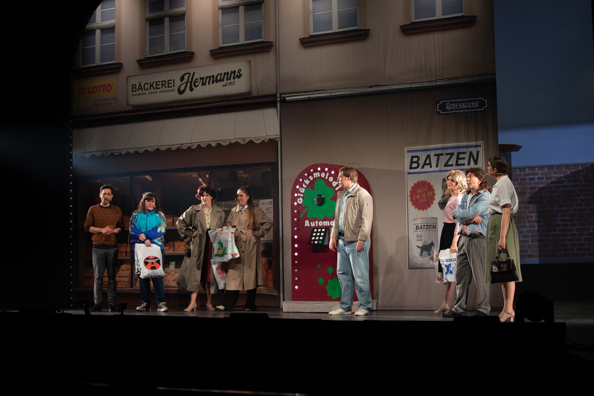 Eine Gruppe von Menschen steht auf einer Bühne, die einer Bäckerei nachempfunden ist. Einige haben Einkaufstüten in der Hand, andere scheinen sich zu unterhalten. In der Mitte steht eine rote Glücksmaschine.
