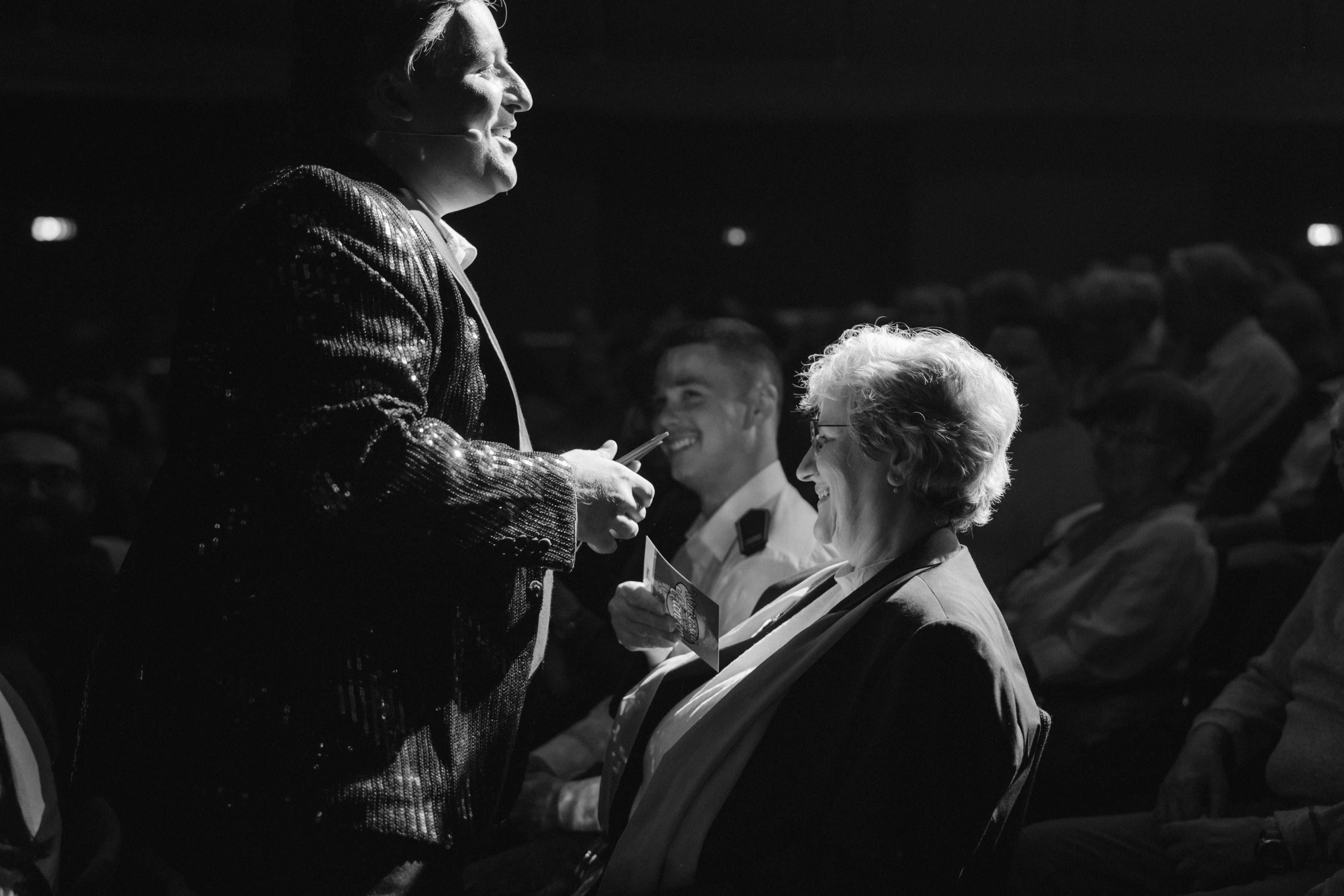 Ein Mann im glitzernden Sakko geht lächelnd durch die Publikumsreihe und spricht eine sitzende Zuschauerin an, umgeben von weiteren lachenden Personen.