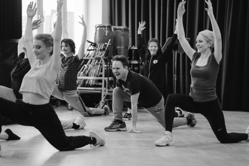 Lachende Person in der Mitte eines Proberaums kniet in sportlicher Pose auf dem Boden, umgeben von tanzenden Menschen mit erhobenen Armen.