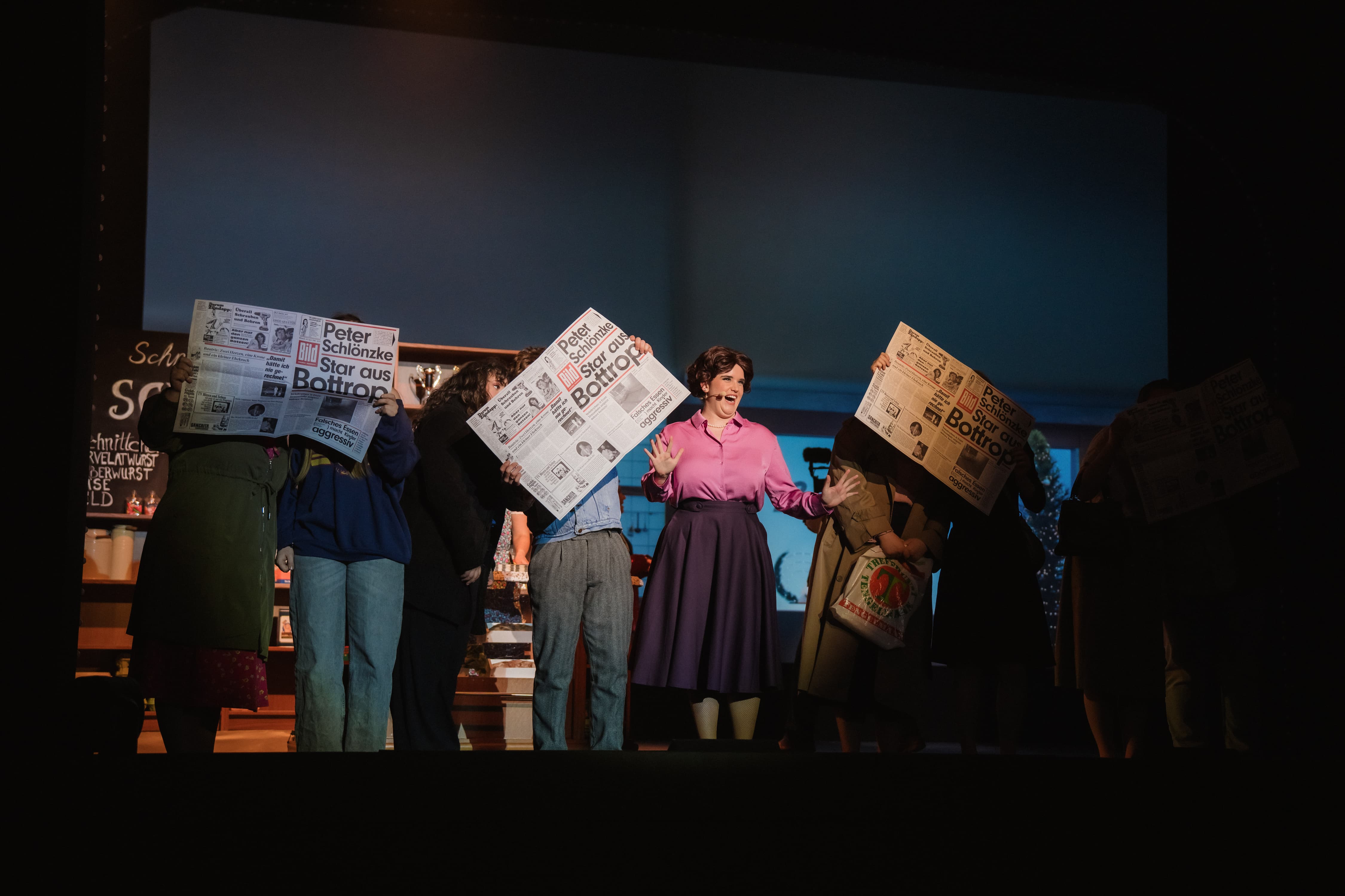 Bühnenszene mit mehreren Personen, fünf davon halten Zeitungen mit der Schlagzeile „Peter Schlönzke – Star aus Bottrop“, eine Frau in pinkem Blusenoutfit singt zentral.