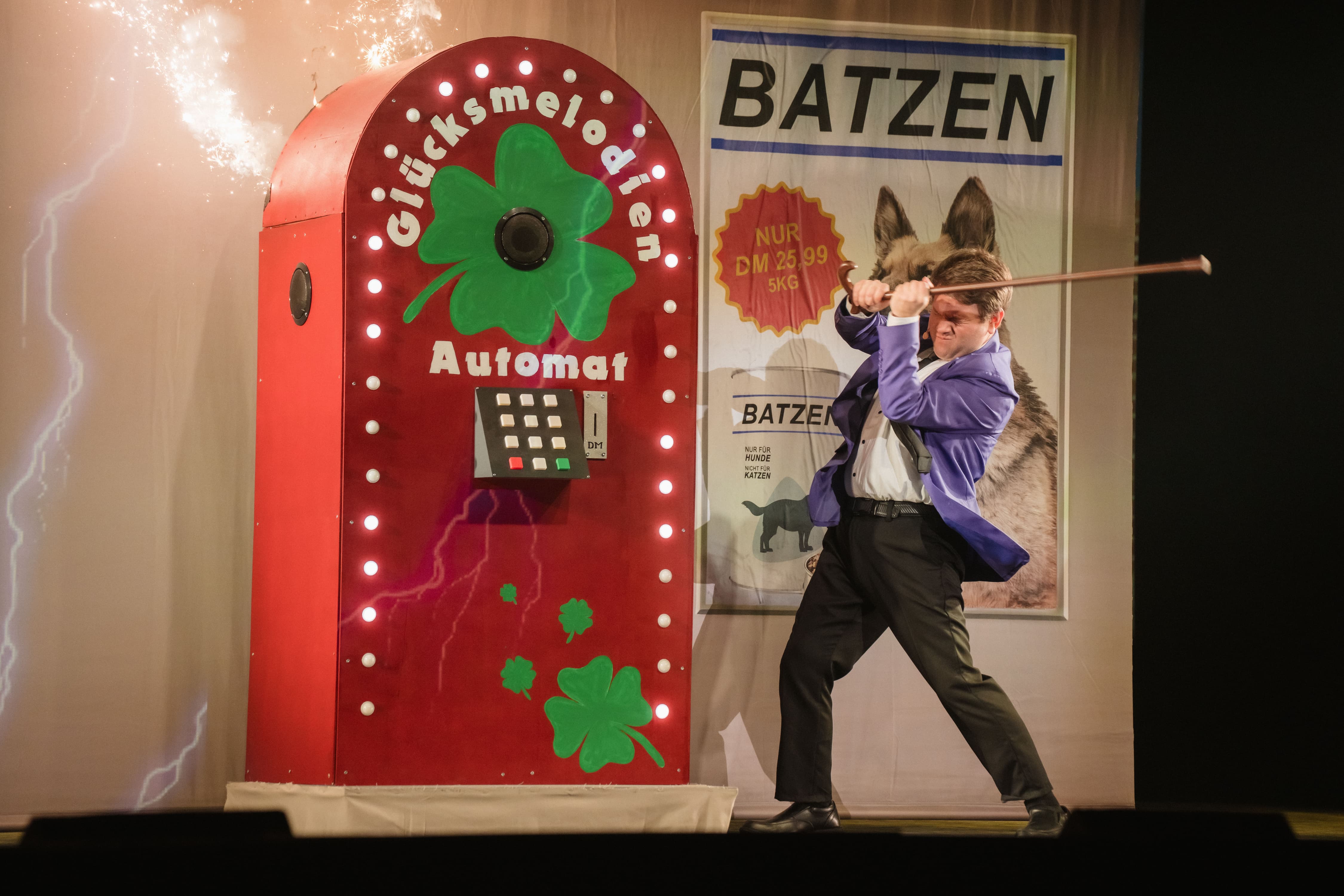 Ein Mann in Anzug schlägt mit einem Stock auf einen funkenstiebenden roten „Glücksmelodien-Automat“, daneben eine große Werbewand mit Hundemotiv.