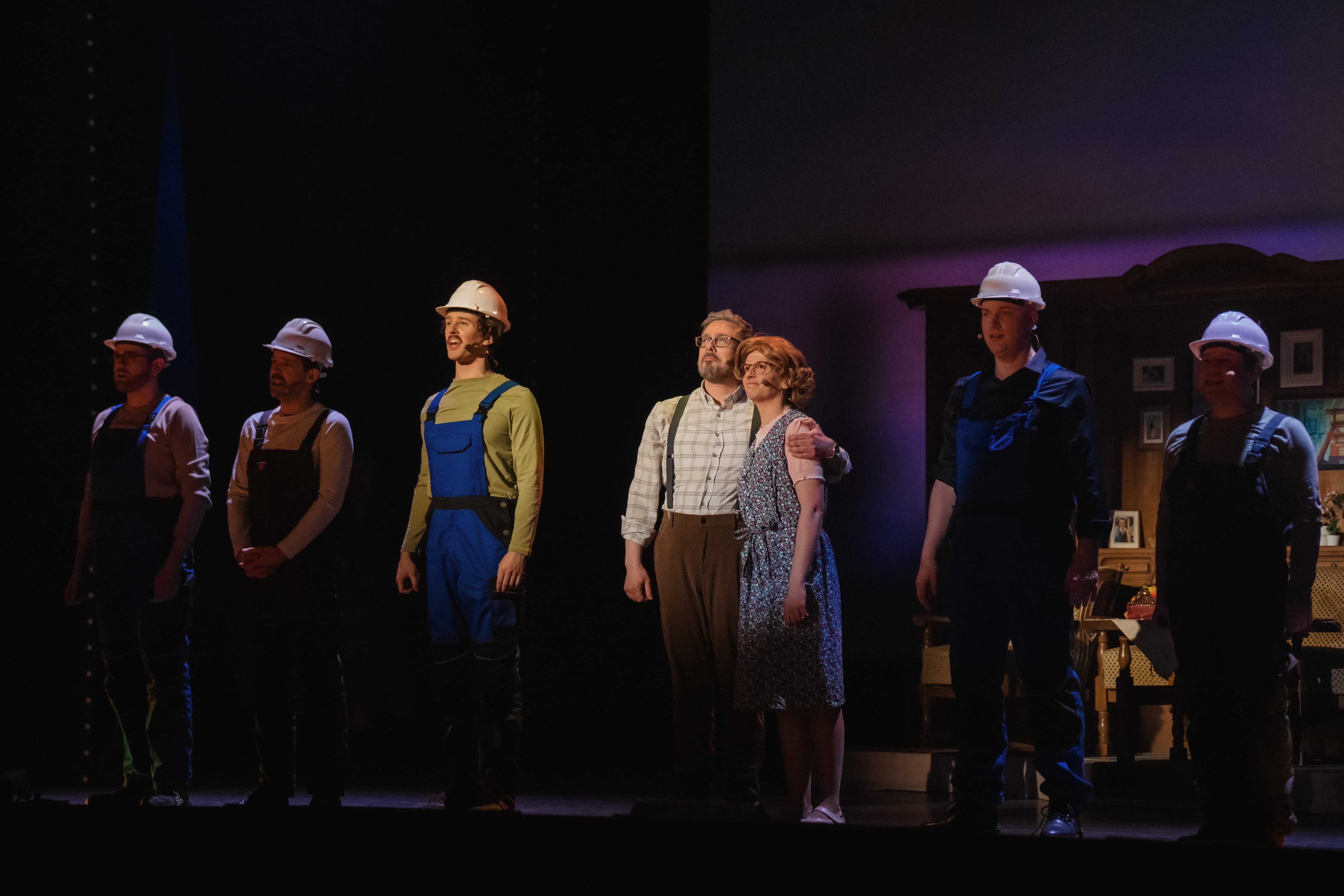 Sieben Personen auf einer Bühne, teils in Bauarbeiterkleidung mit Helmen, zwei in Alltagskostümen, vor einem wohnlichen Bühnenbild mit Schrank.