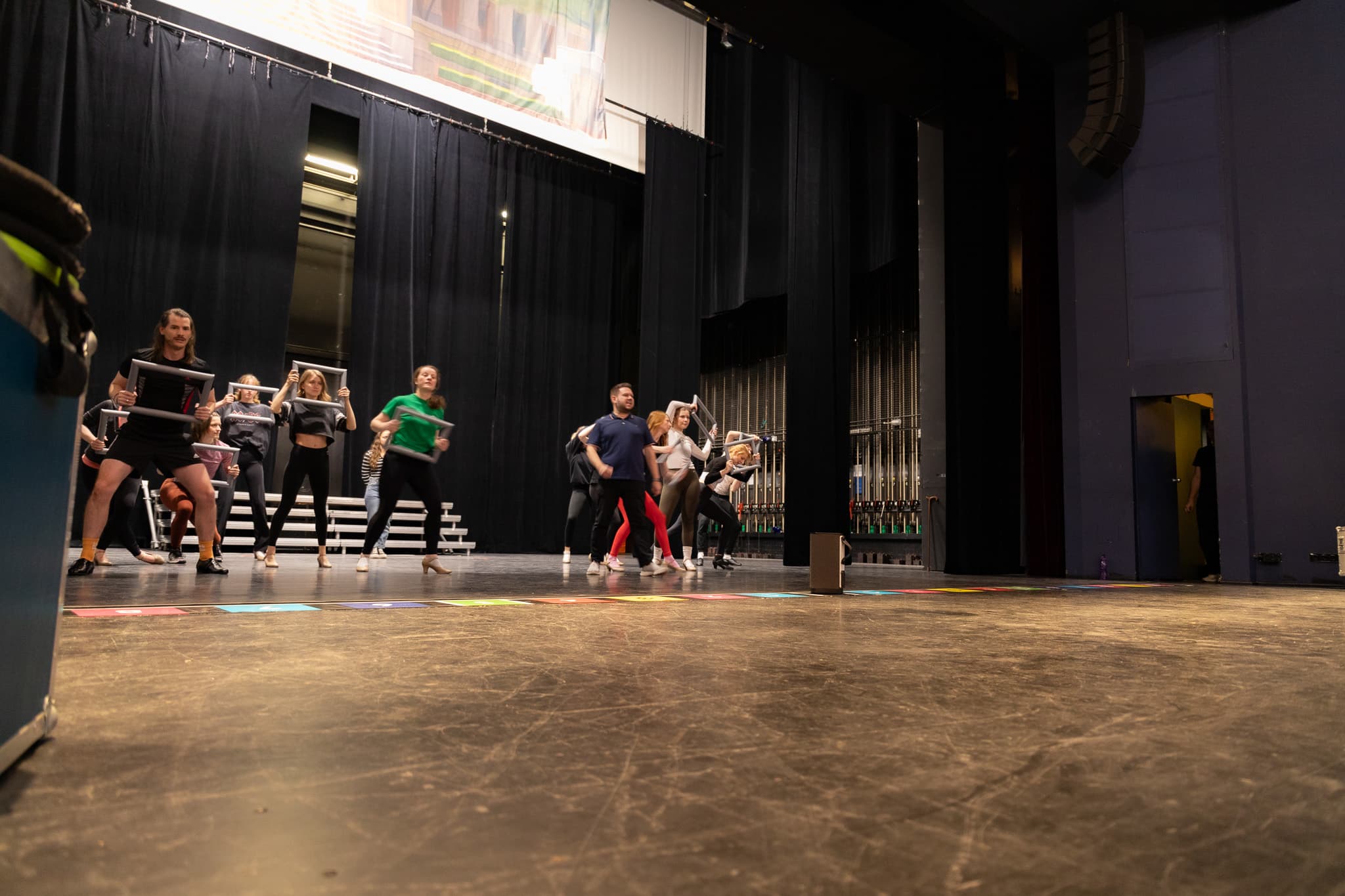 Eine Gruppe von Darstellern probt auf einer Bühne, einige halten silberne Rahmen, vor einem dunklen Bühnenvorhang.